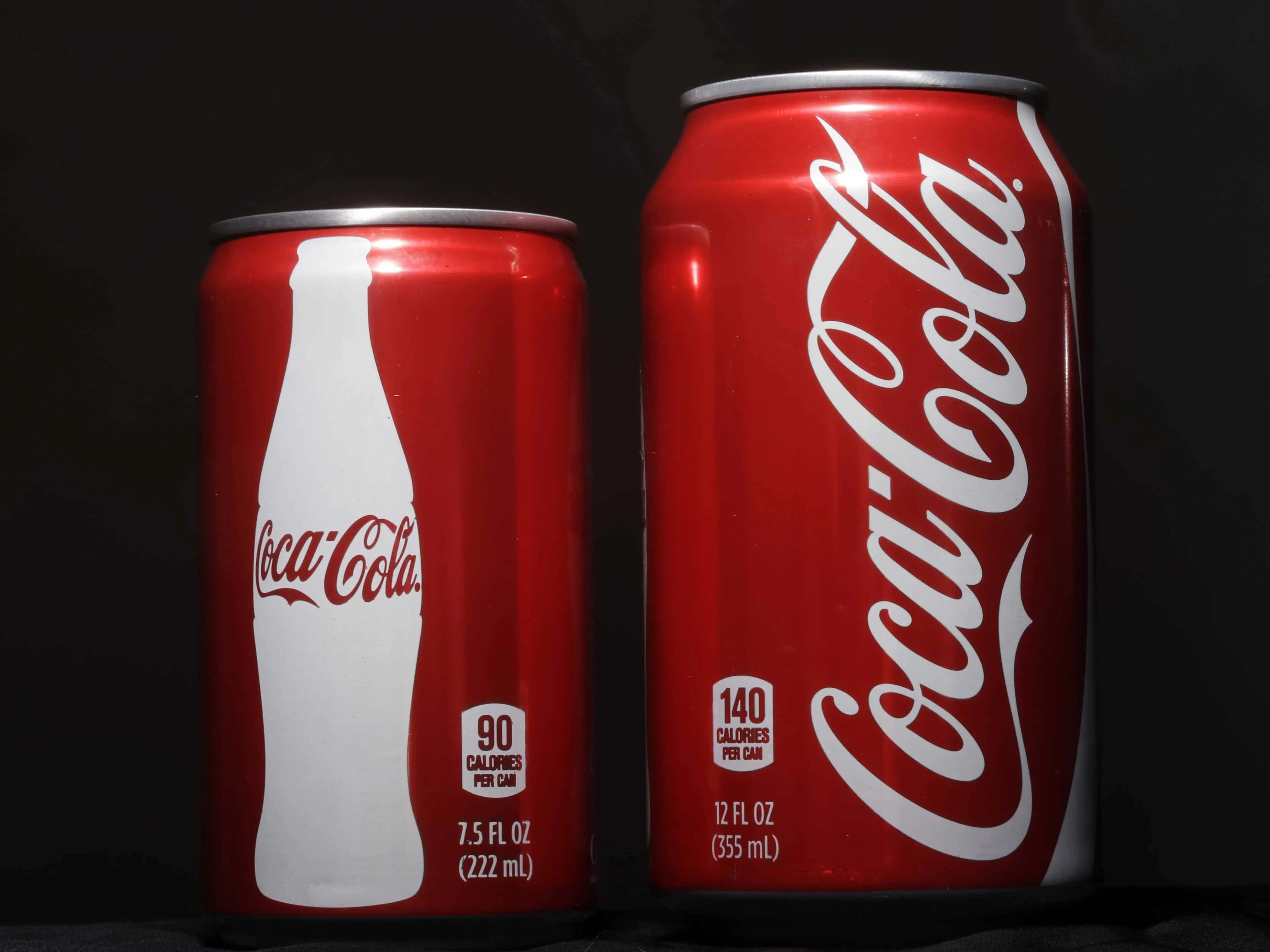 coca cola 桌布 壁紙 — 兩個紅色可口可樂罐，一個小罐有白色瓶形標誌，另一個大罐有草書標誌，在深色背景前。
