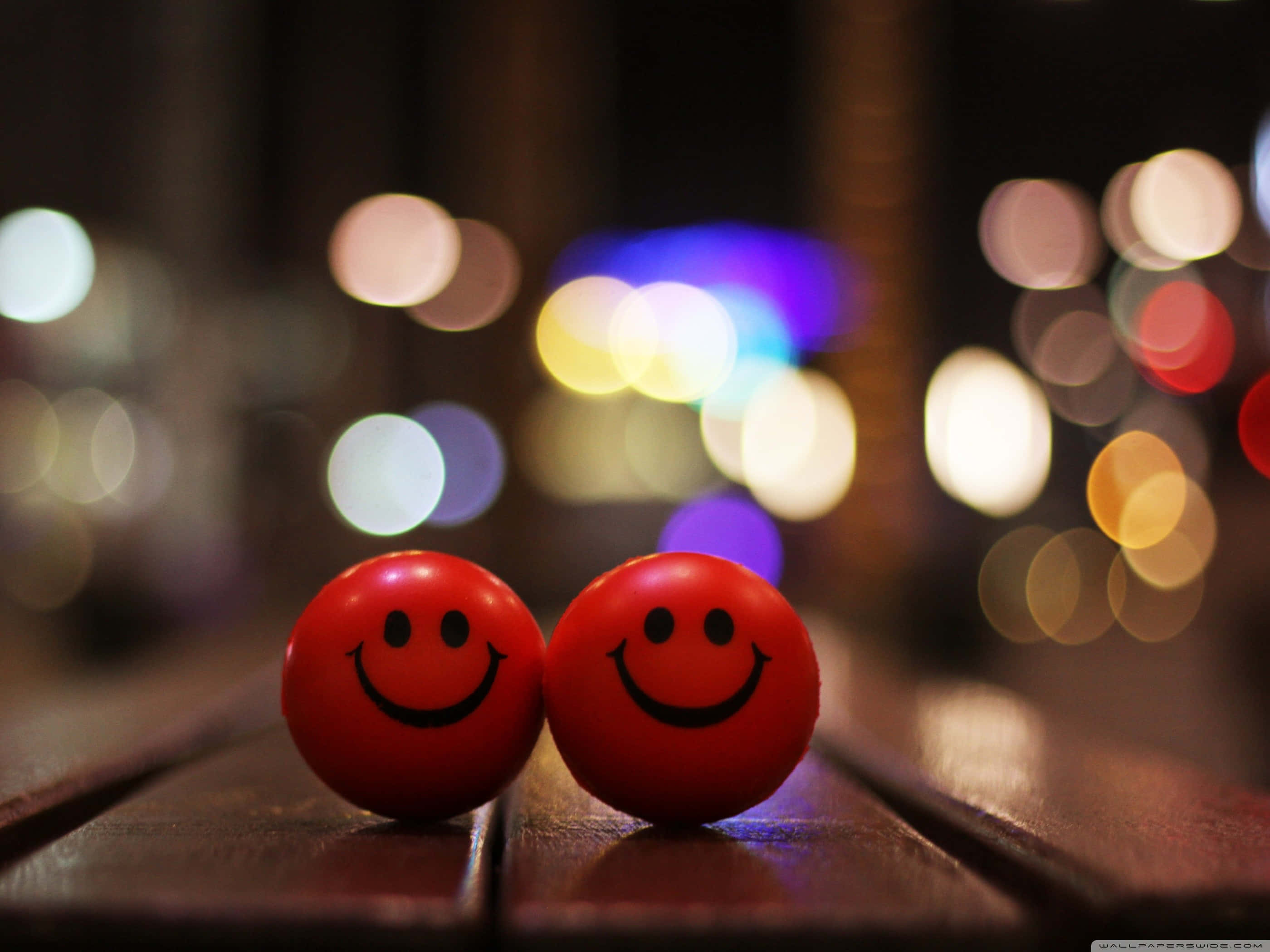 cute happy 桌布 壁紙 — 兩個紅色笑臉球並排放在木質表面上，背景是夜晚中柔和發光的彩色散景燈光。