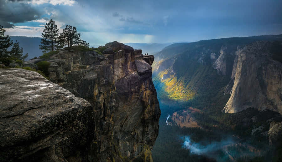 Cool Landscape 桌布 壁紙 — 兩個細小的人影站在崎嶇的懸崖邊緣，俯瞰著一個深谷，谷中有秋天的樹木和霧氣，上方是戲劇性的多雲天空。