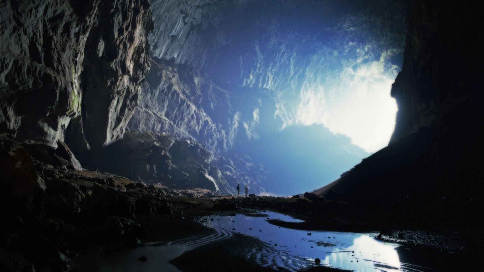 Cave 桌布 壁紙 — 兩個細小的人物站在巨大洞穴的洞口，敬畏地望著，陽光從上方湧入，照亮了岩石牆壁和下方反射的溪流。