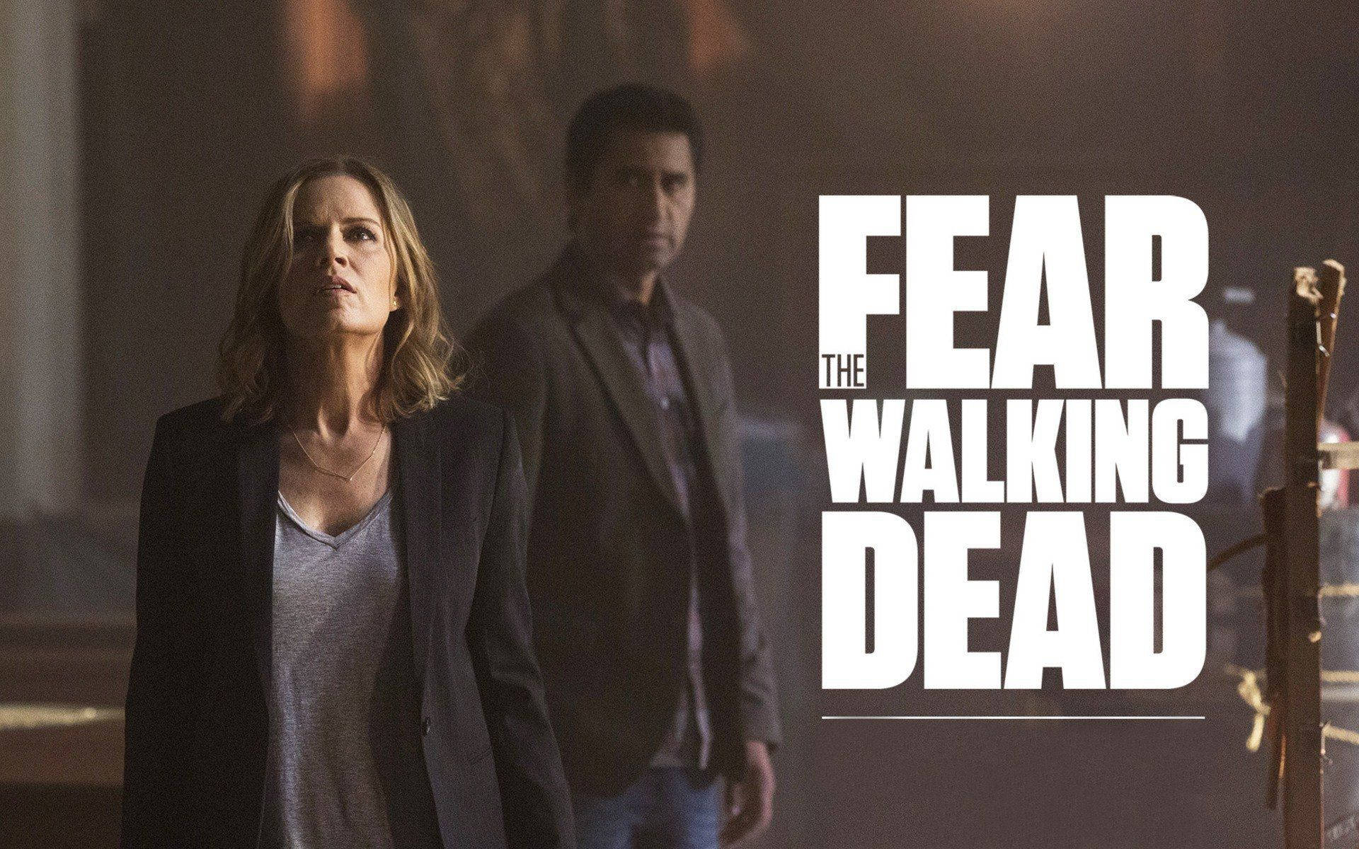 dead 桌布 壁紙 — 兩個緊張的角色站在燈光昏暗的房間裡，標題「The Fear The Walking Dead」以粗體白色大字呈現。