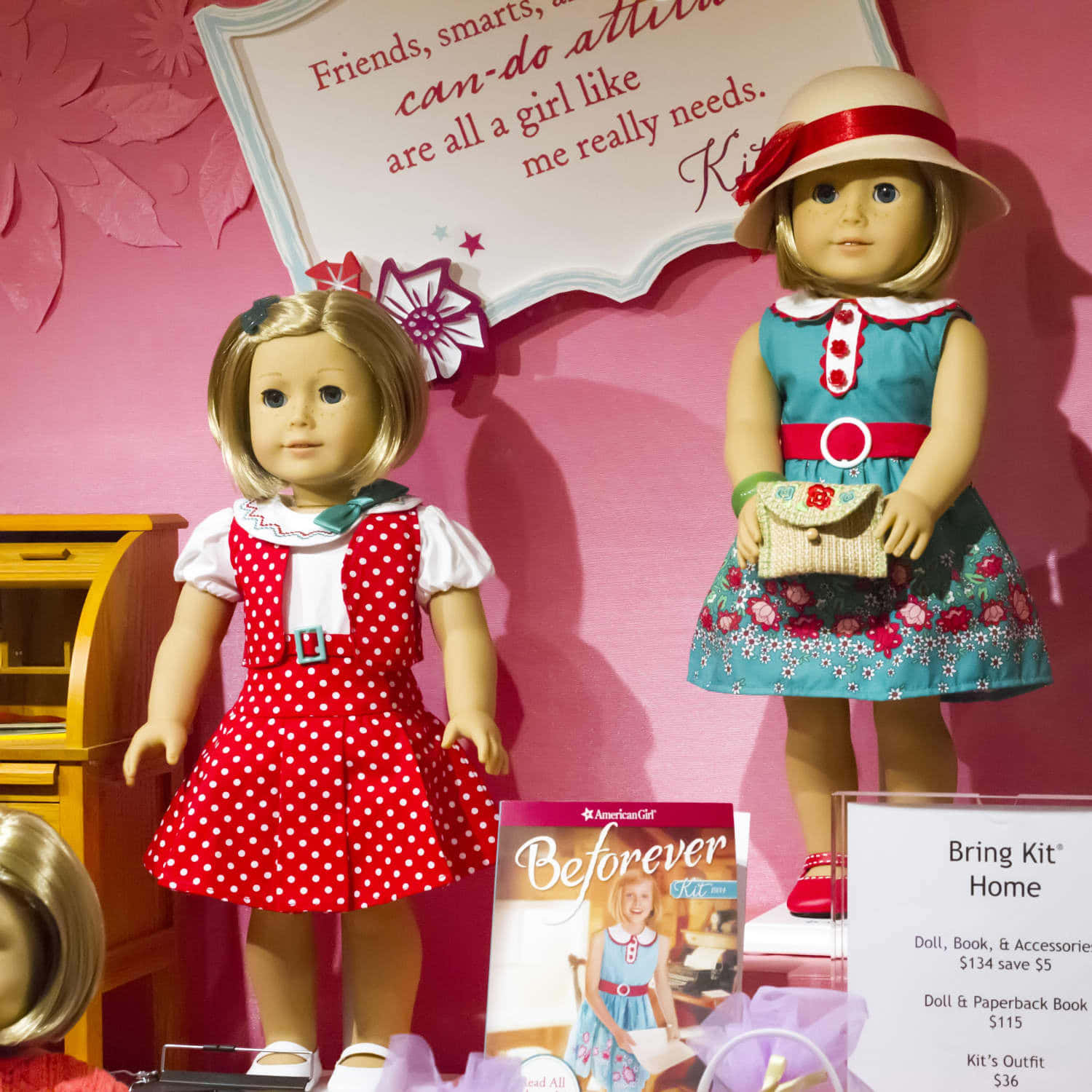american girl 桌布 壁紙 — 兩個美國女孩娃娃穿著色彩繽紛的服裝，站在粉紅色花卉背景前的宣傳材料旁邊。