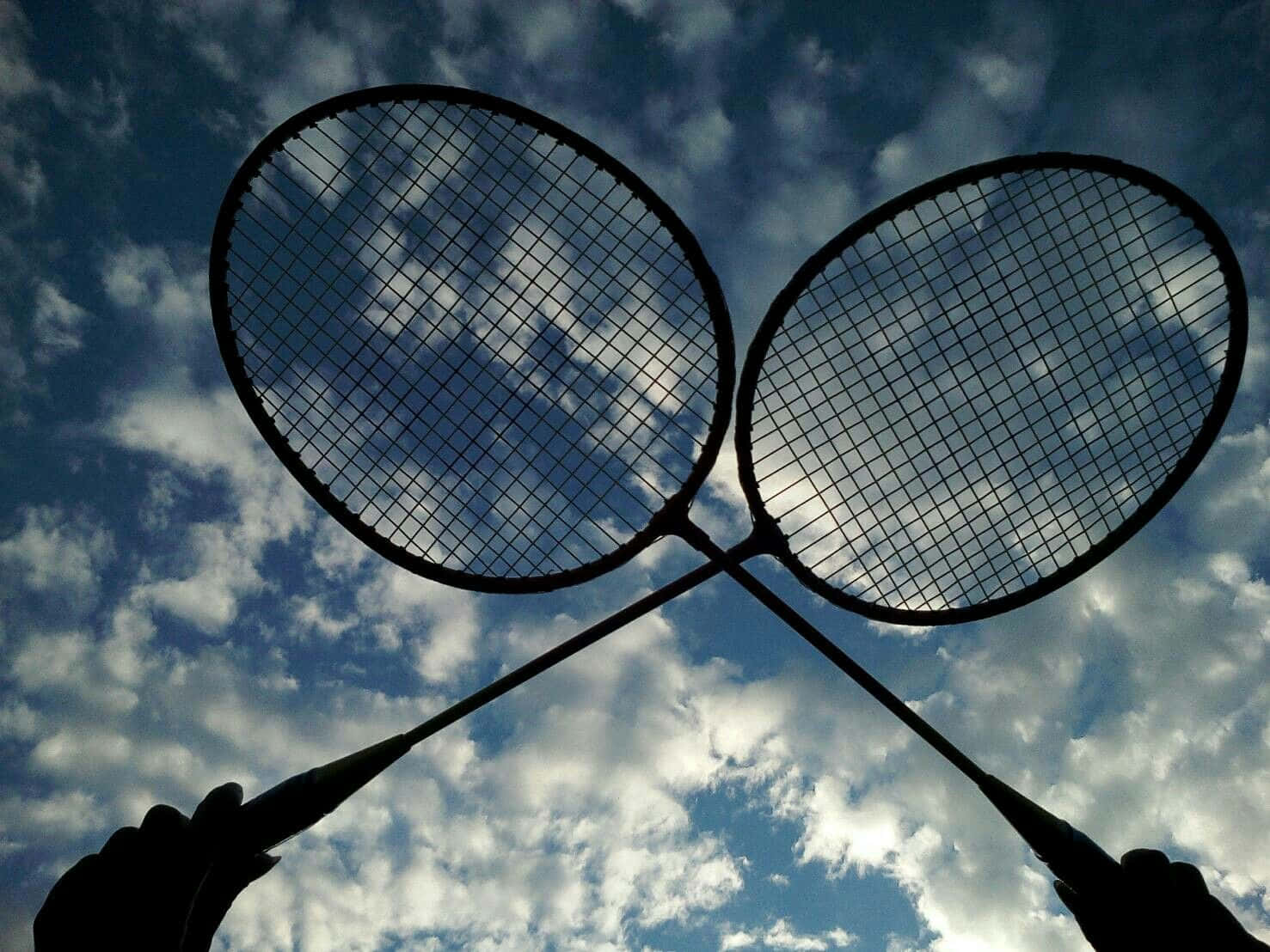 HD Badminton 桌布 壁紙 — 兩個羽毛球拍剪影，背景是戲劇性的多雲天空，底部由手握住。