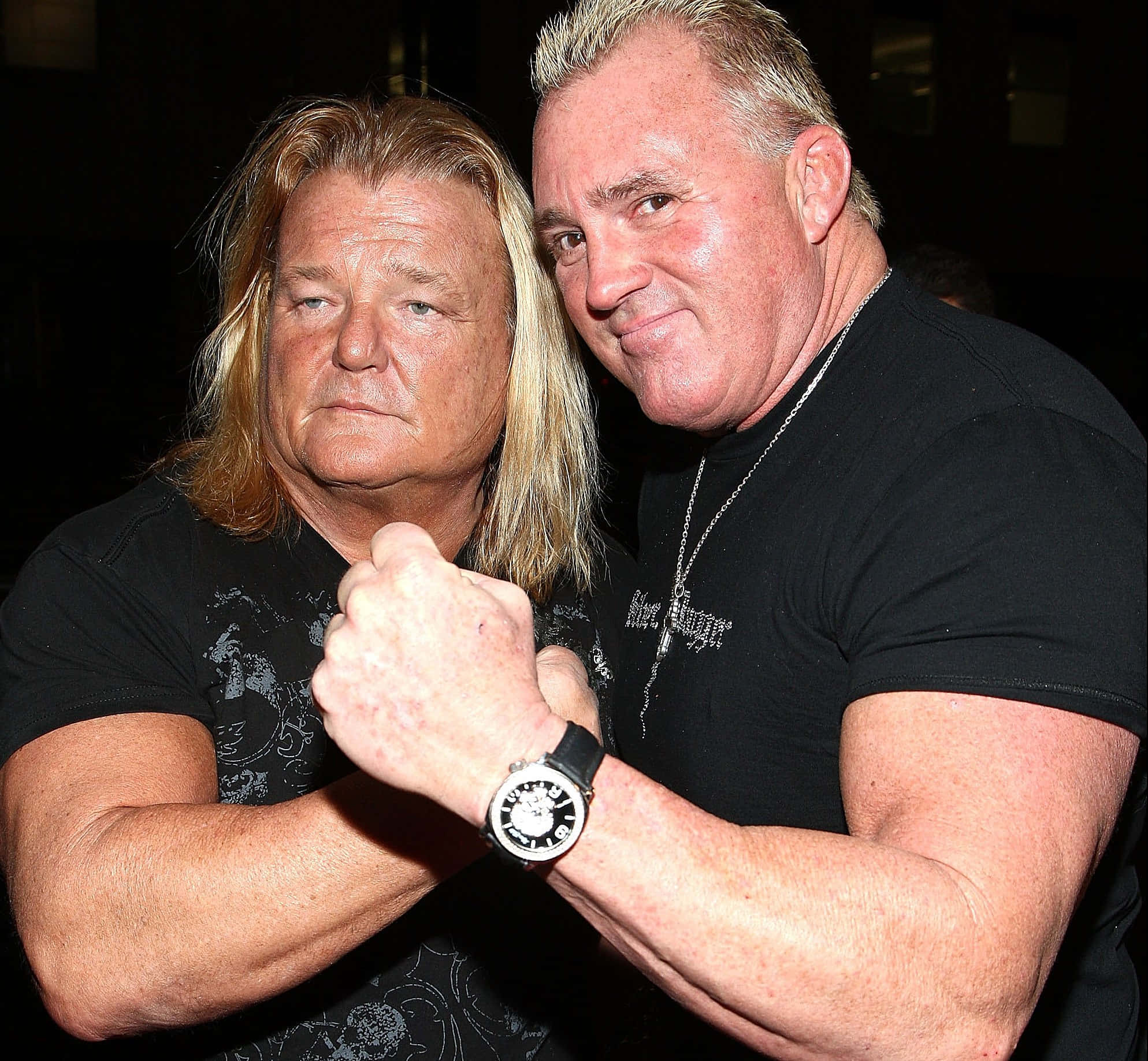 WWE 桌布 壁紙 — 兩個肌肉發達的男人穿著黑色T恤，在昏暗燈光下緊密地擺姿勢，其中一個用拳頭撞擊另一個的手臂。