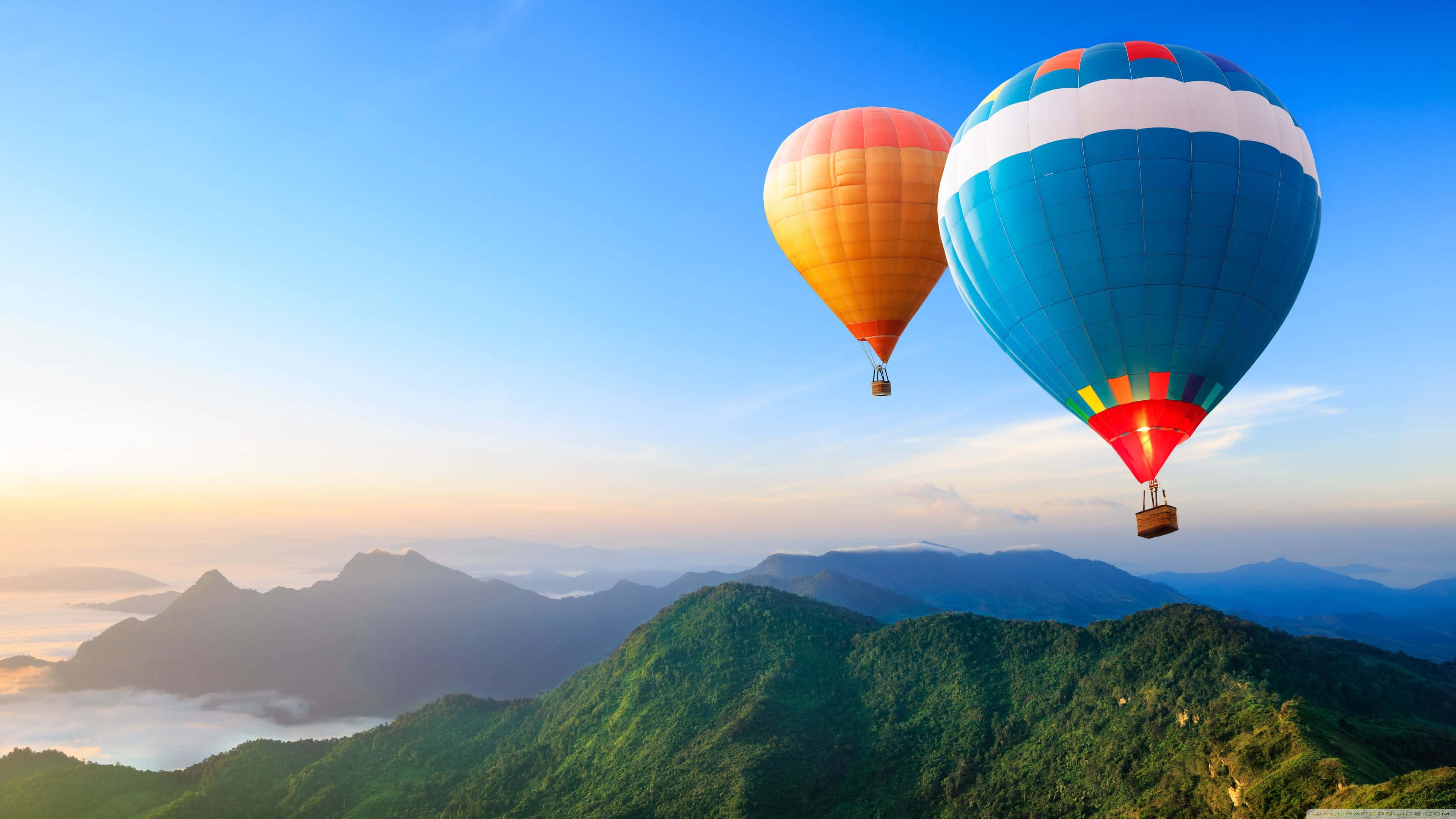 Best Travel 桌布 壁紙 — 兩個色彩繽紛的熱氣球在日出時飄浮在鬱鬱蔥蔥的綠色山脈之上，下方有薄霧。