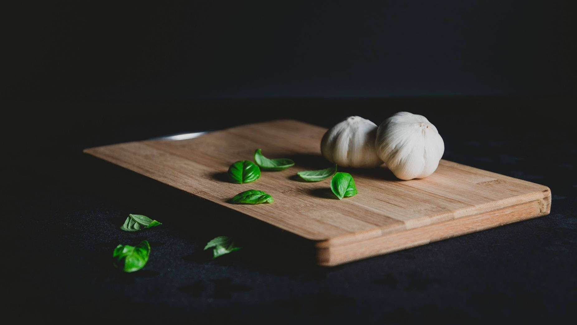 Garlic 桌布 壁紙 — 兩個蒜頭和新鮮羅勒葉放在木製砧板上，背景為深色。