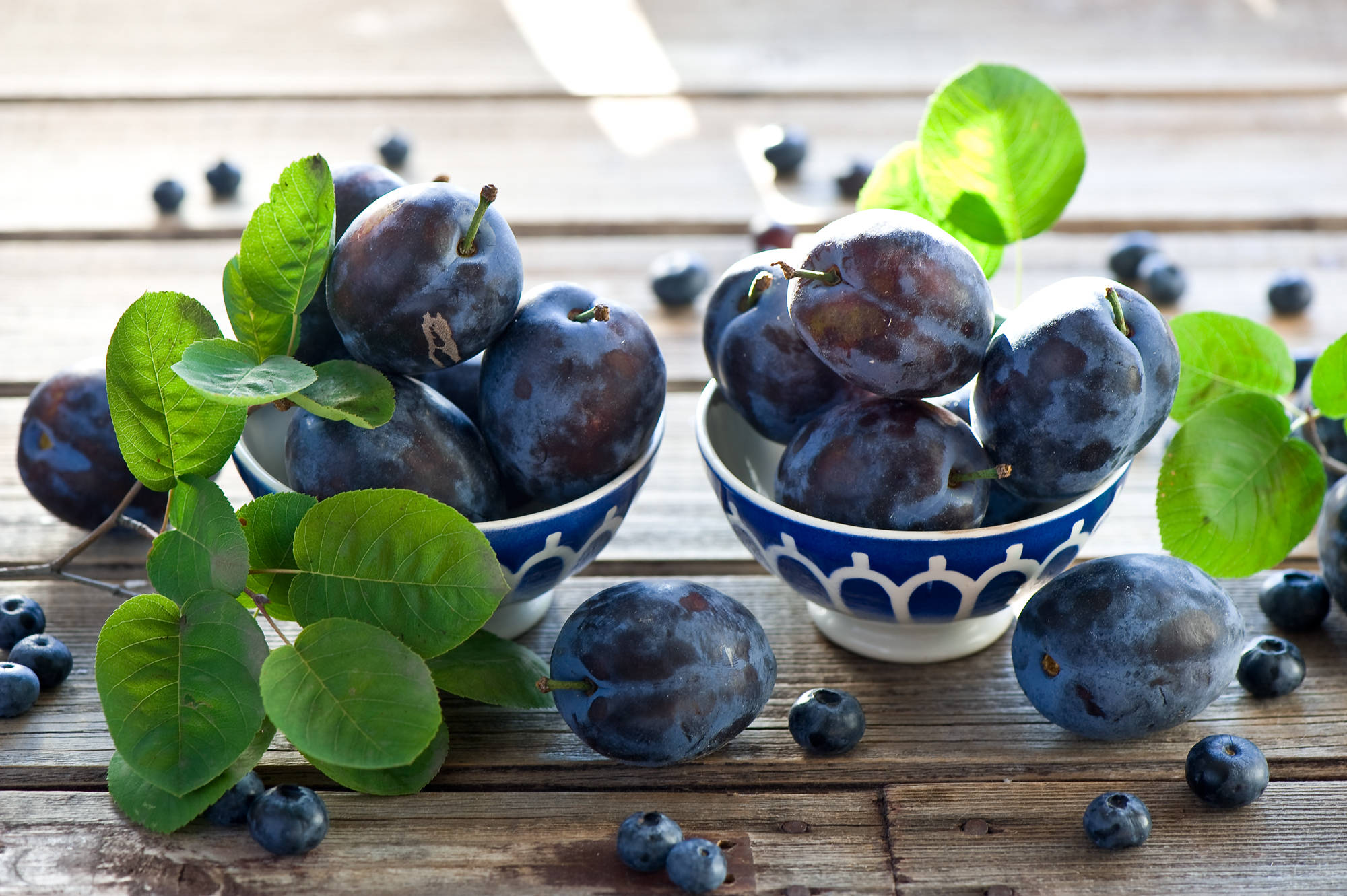 Fruits 桌布 壁紙 — 兩個藍白圖案碗裝滿了李子和藍莓，放在風化的木桌上，配上新鮮的綠葉。