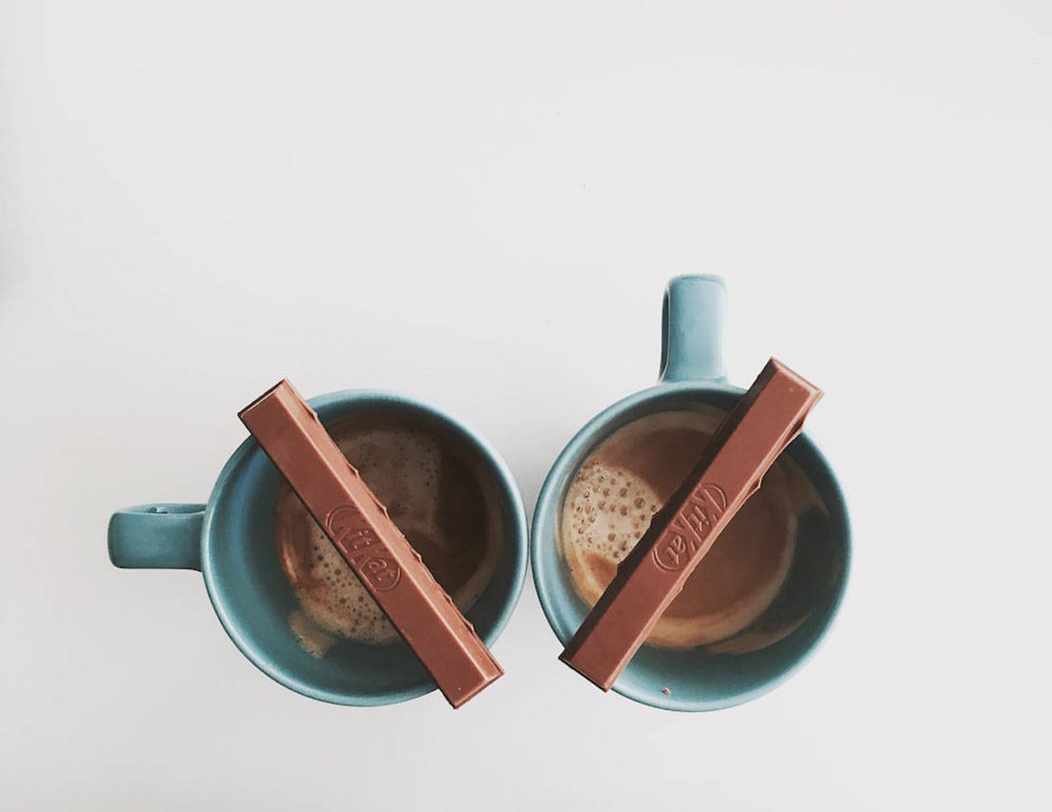 Chocolate 桌布 壁紙 — 兩個藍綠色馬克杯裝滿泡沫熱巧克力，每個杯緣斜放著一條KitKat巧克力棒。