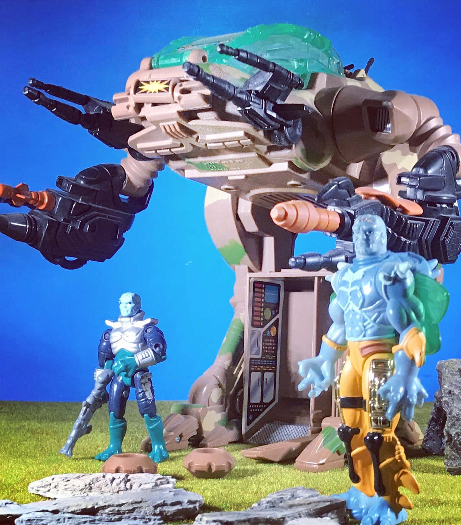 toys 桌布 壁紙 — 兩個藍色皮膚的動作人偶站在一個大型棕綠色裝甲機甲前面，有多個大砲，背景是明亮的藍天。