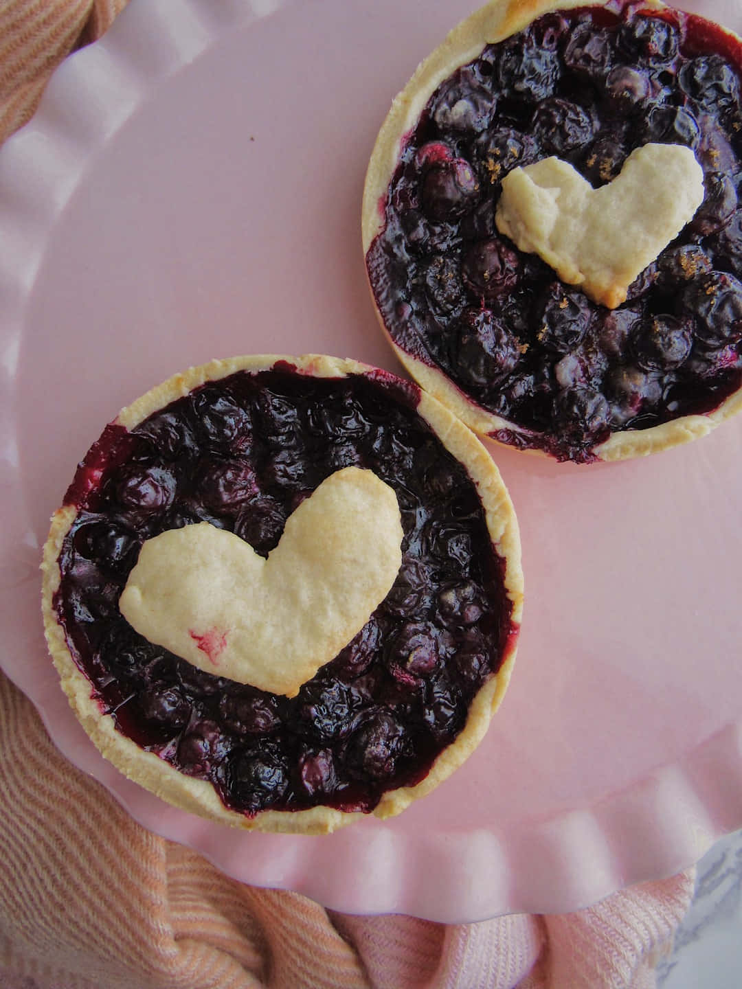 Blueberries Tart 桌布 壁紙 — 兩個藍莓派，上面有愛心形狀的酥皮，放在粉紅色盤子上，盤子下墊著蜜桃色布料。