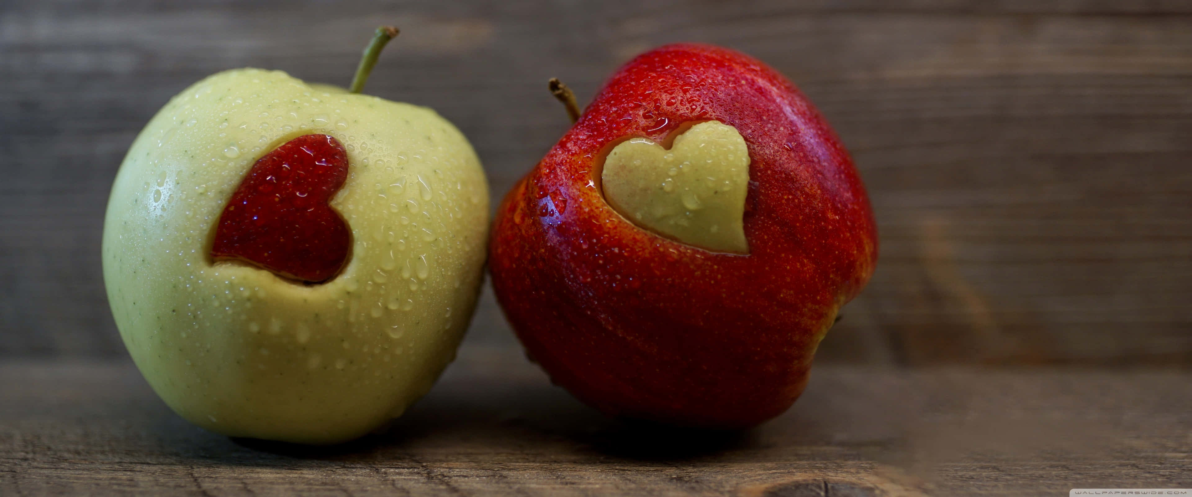 Apple 3840x1600 桌布 壁紙 — 兩個蘋果，一個綠色一個紅色，每個都有一個心形咬痕，露出對方的果肉，放在木質表面上。