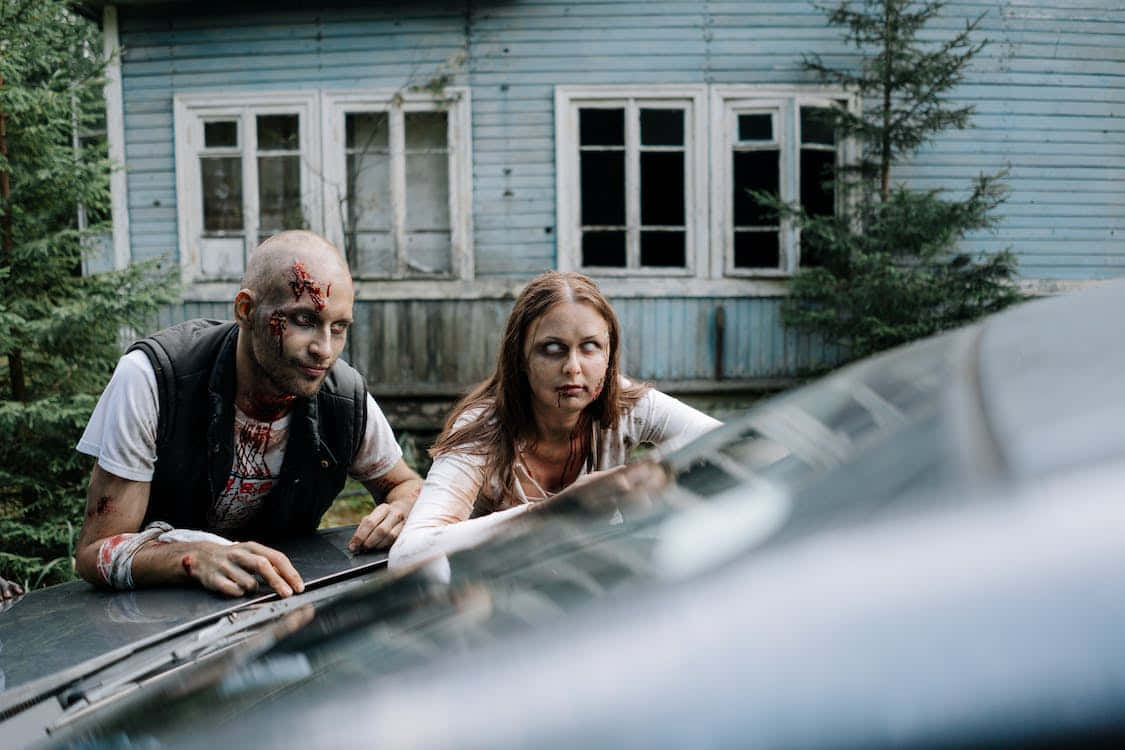 Scary Zombie 桌布 壁紙 — 兩個血淋淋的殭屍靠在一輛汽車的引擎蓋上，前面是一間破舊的藍色房子，周圍是常青樹。