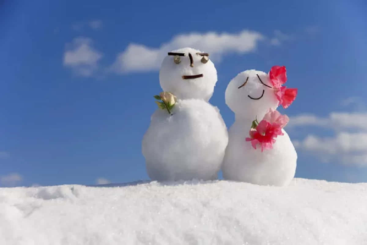 冬天 桌布 壁紙 — 兩個表情豐富、帶有粉紅色花朵的雪人並肩站在白雪覆蓋的山丘上，背景是明亮的藍天。