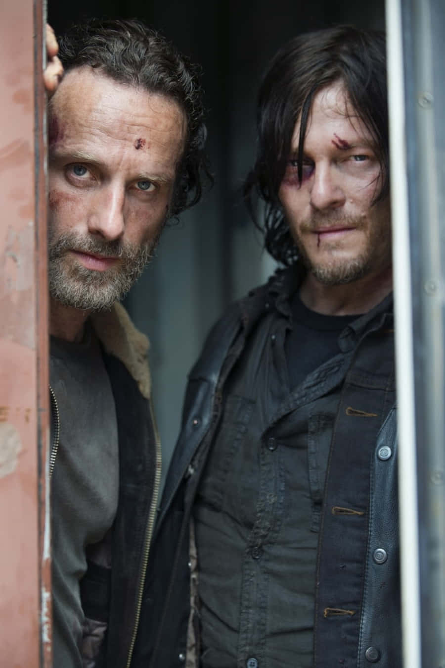walking dead 桌布 壁紙 — 兩個被打得遍體鱗傷、滿臉是血的男人，在一個粗糙的城市環境中，透過一扇生鏽的金屬門窺視。