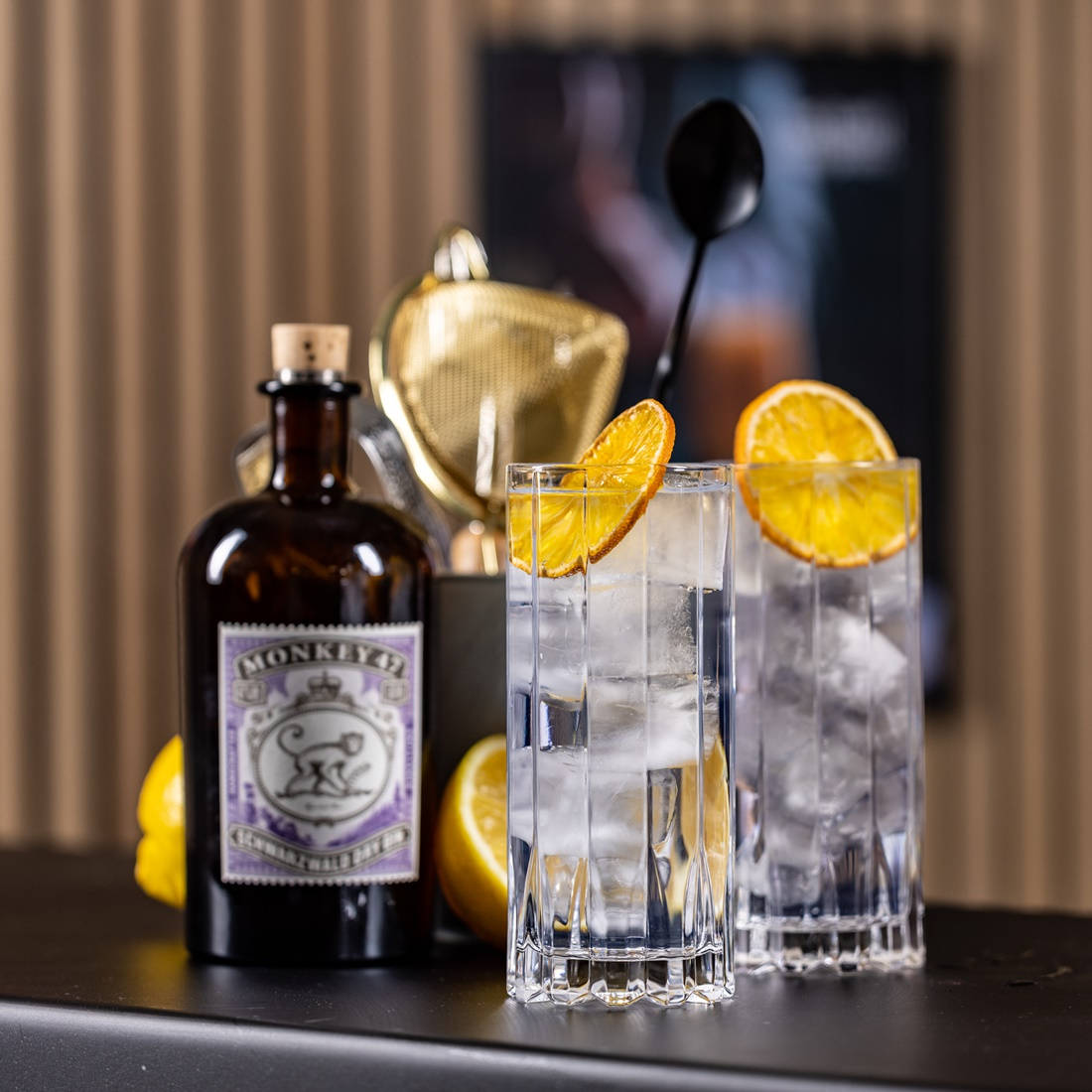 cocktail 桌布 壁紙 — 兩個裝滿冰塊和橙片的水晶杯放在一個光滑的黑色吧檯上，旁邊是一瓶深色的Monkey 47琴酒。