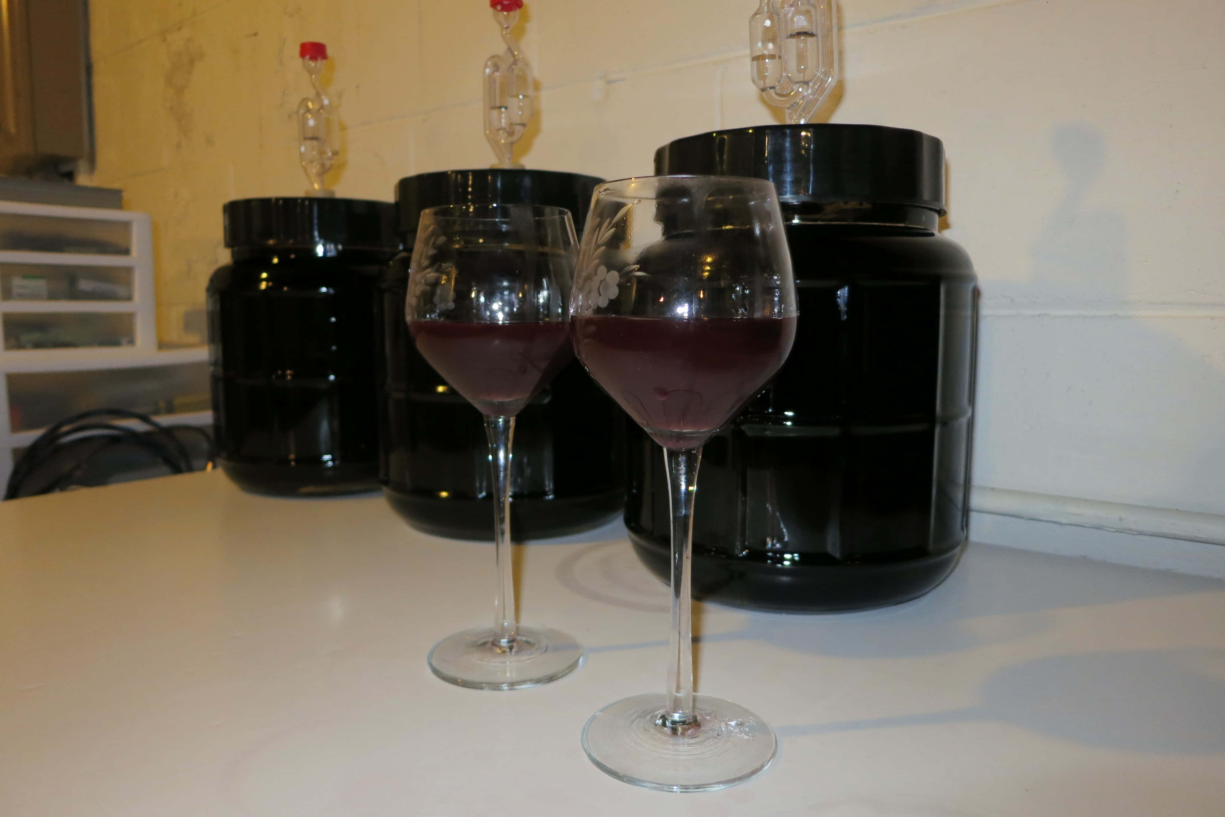 Blackberry Wine 桌布 壁紙 — 兩個裝滿深紅色液體的酒杯放在白色檯面上，前面是帶有空氣鎖的黑色發酵桶。