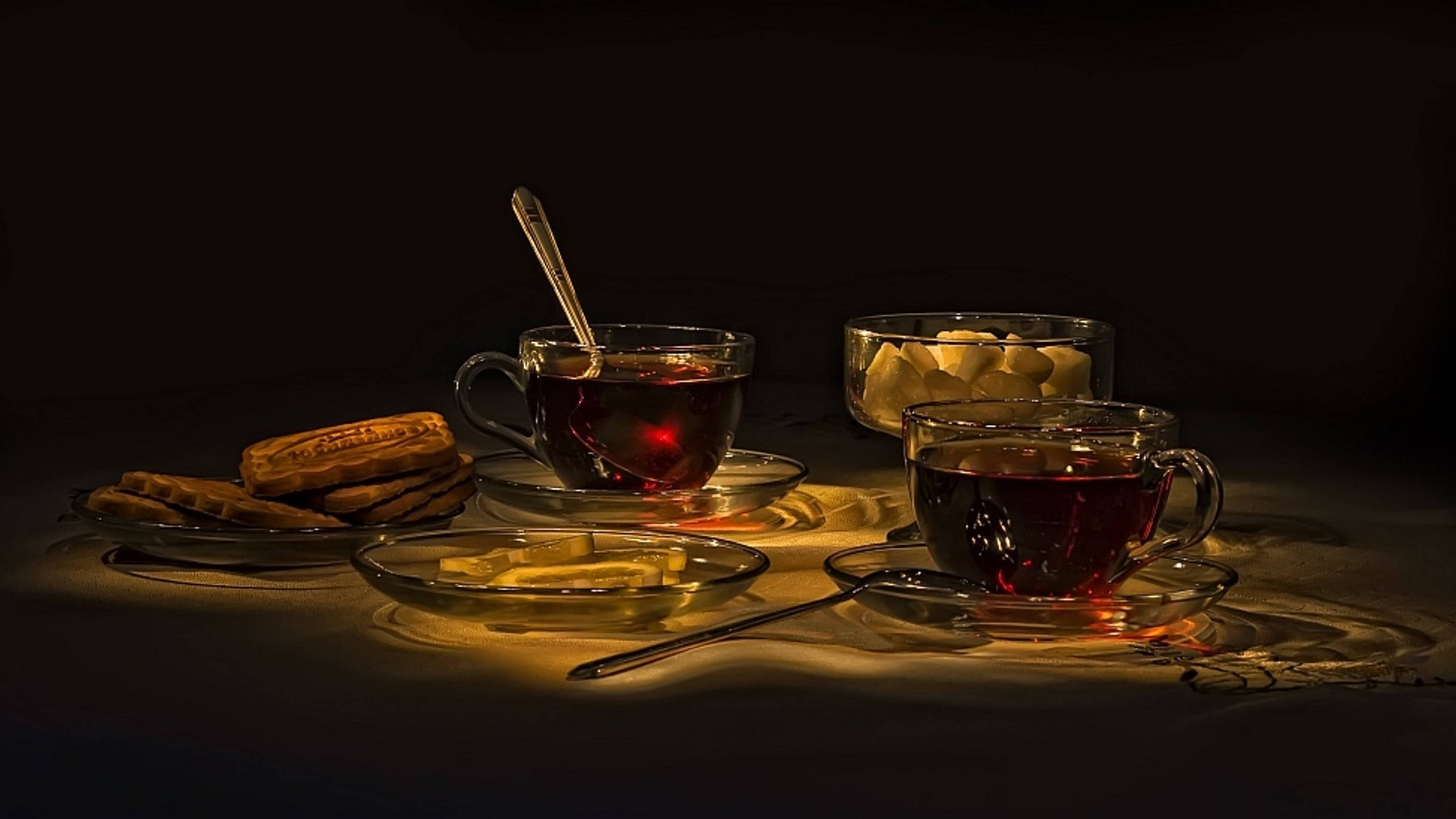 tea 桌布 壁紙 — 兩個裝滿深色茶的玻璃茶杯在聚光燈下溫暖地發光，旁邊是餅乾和方糖，放在深色桌子上。