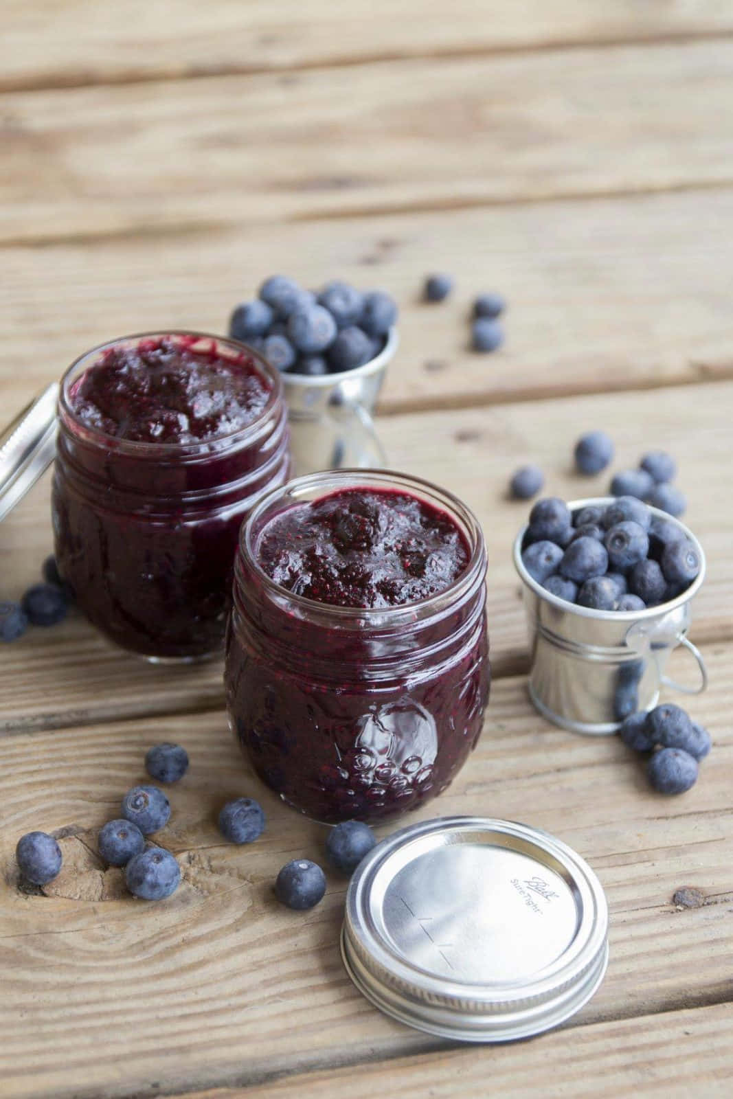 Blueberry Jam 桌布 壁紙 — 兩個裝滿閃亮藍莓醬的玻璃罐放在一張風化的木桌上，周圍是新鮮藍莓和小金屬桶。