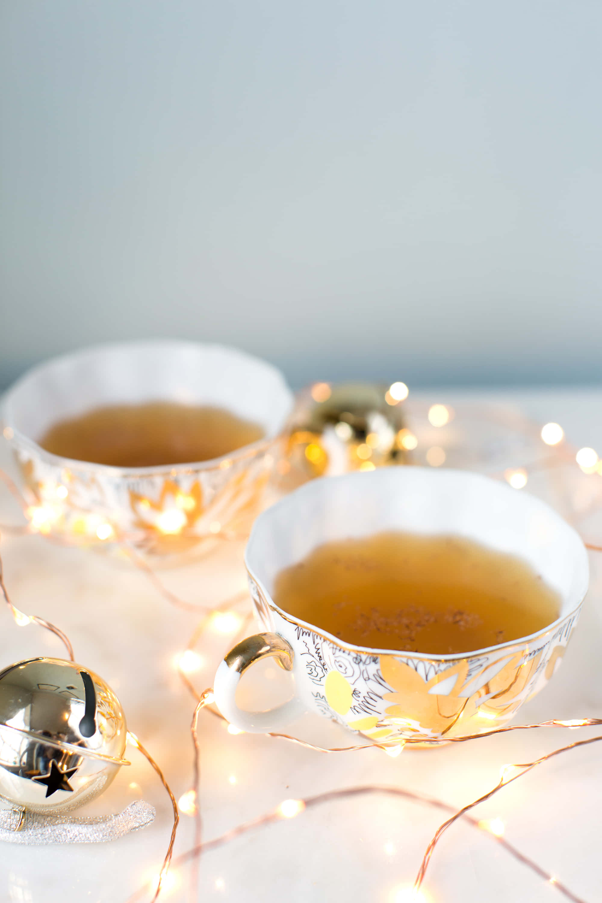 Winter Drinks 桌布 壁紙 — 兩個裝飾精美的茶杯盛滿金黃色的茶，在柔軟的白色表面上與閃爍的仙女燈和銀鈴一起溫暖地發光。