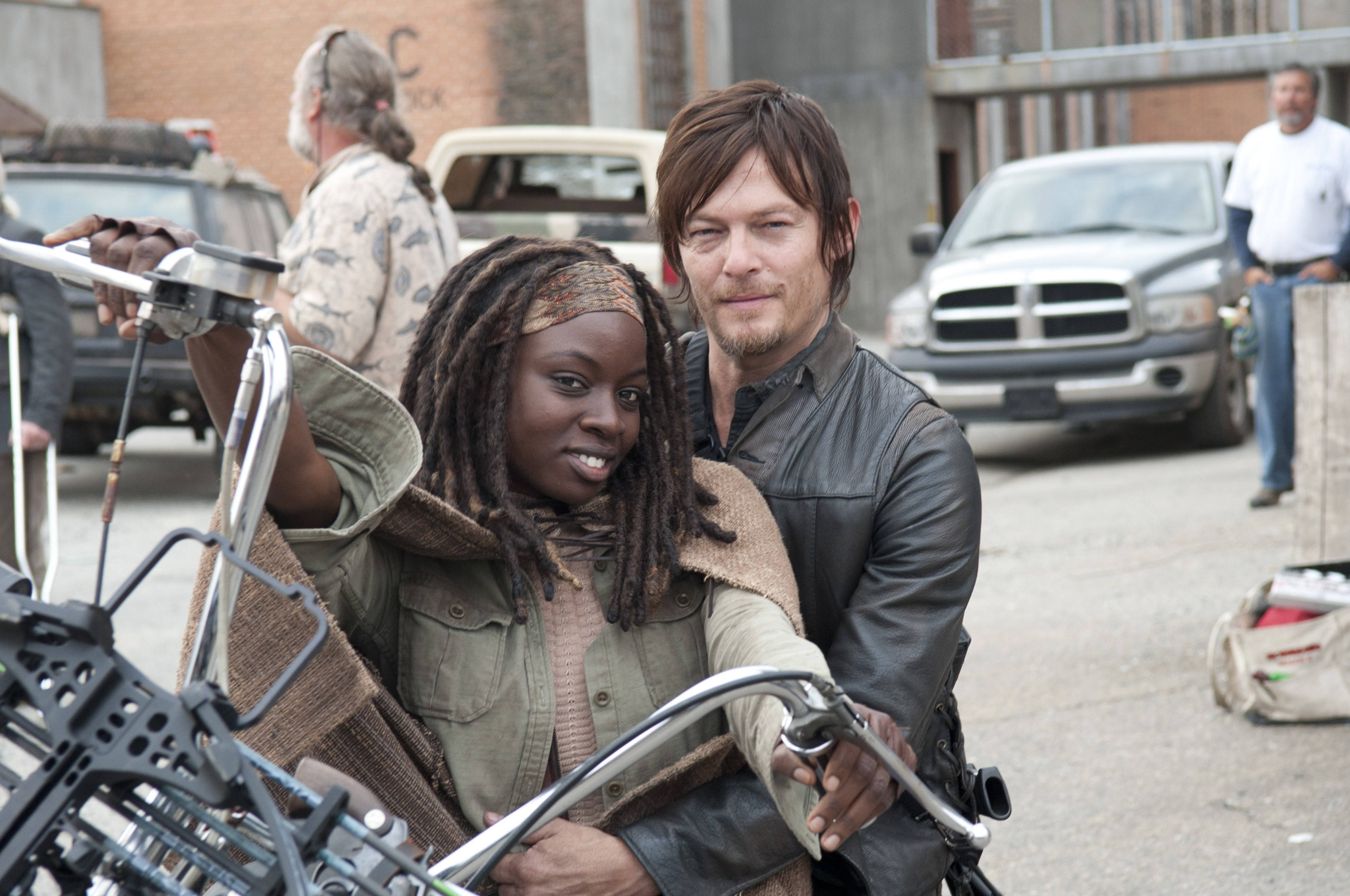 Walking Dead Daryl 桌布 壁紙 — 兩個角色騎著摩托車穿過一個骯髒的城市荒地，背景有廢棄車輛和破舊建築物。