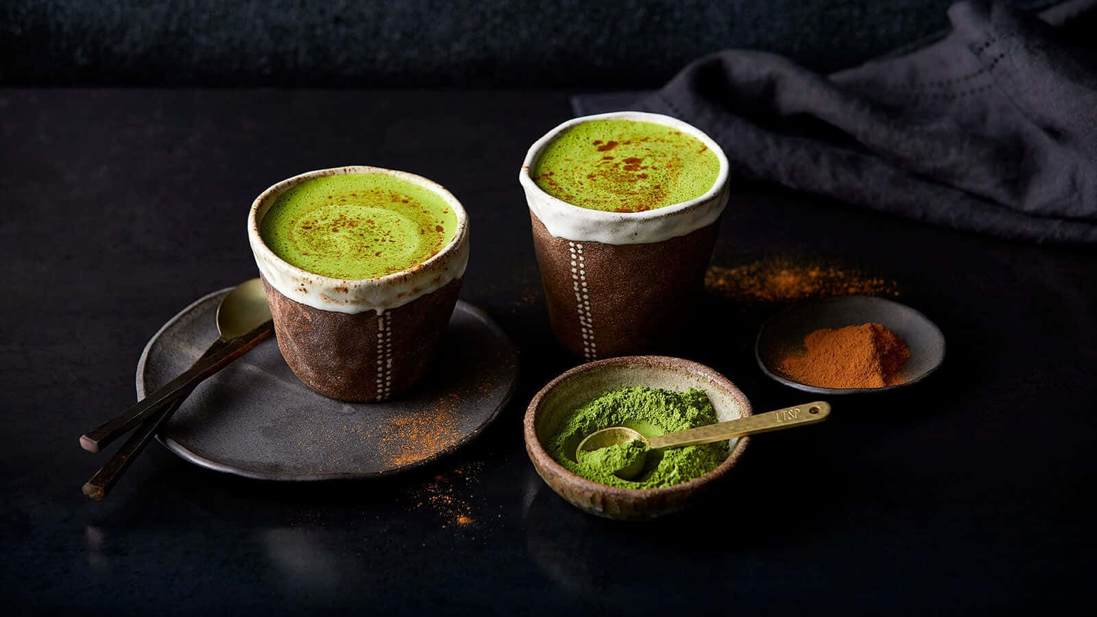 Matcha 桌布 壁紙 — 兩個質樸的陶瓷杯盛載著鮮綠色抹茶，撒上橙味香料，旁邊放著粉末碗，置於深色紋理表面上。