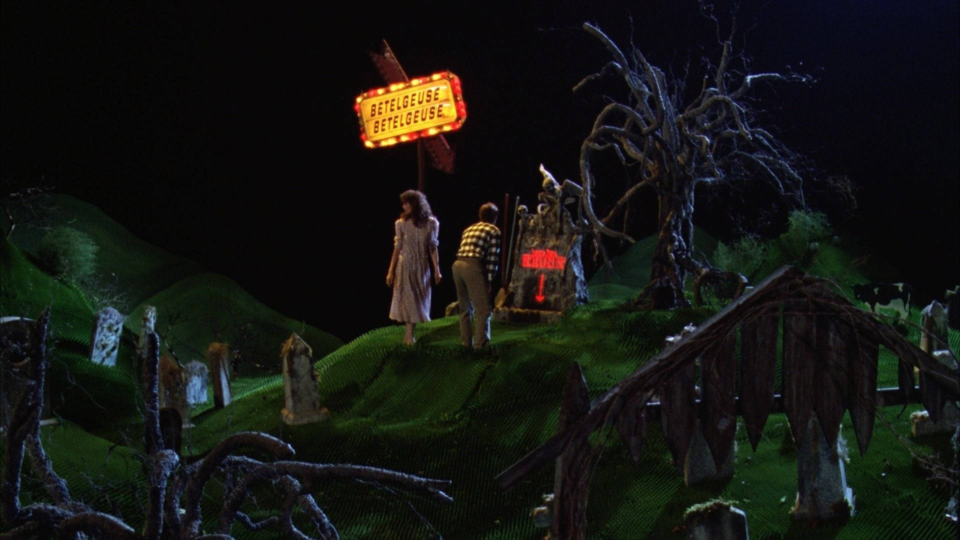 Beetlejuice 桌布 壁紙 — 兩個身影夜晚站在長滿苔蘚的山丘上，在發光的 'BETELGEUSE' 標誌下，附近有一個帶紅光的墓碑。
