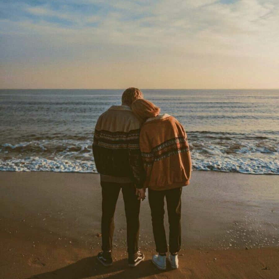Aesthetic Couple 桌布 壁紙 — 兩個身穿圖案外套的人在海灘上日落時分緊靠在一起，眺望著海浪。