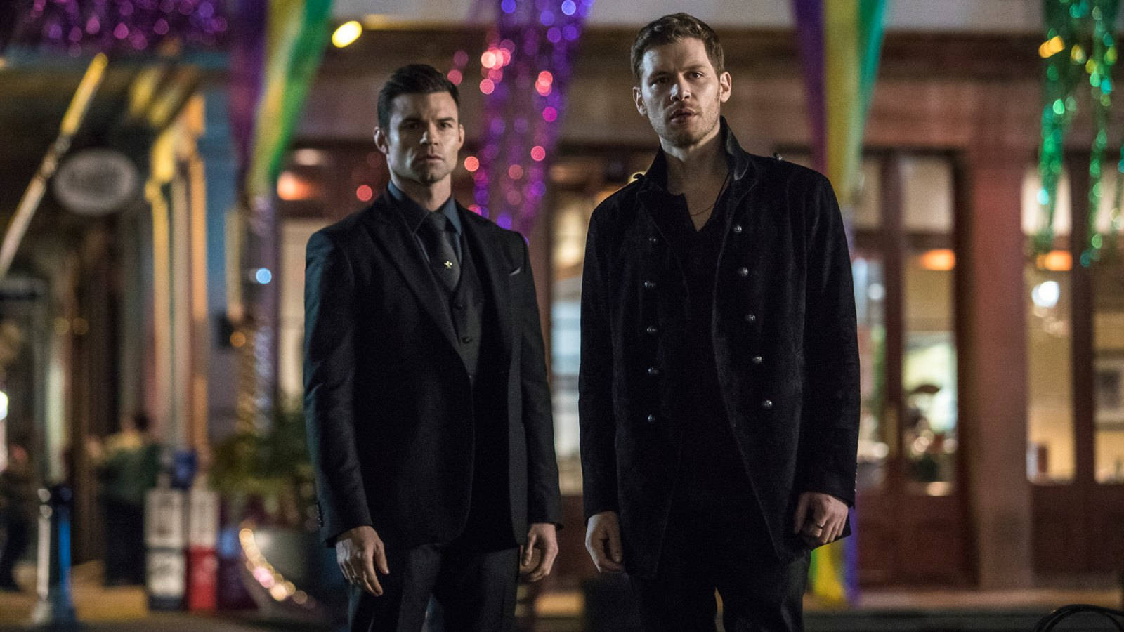 elijah mikaelson 桌布 壁紙 — 兩個身穿深色西裝的男人自信地站在一條充滿活力、裝飾著繽紛節日燈飾的夜間城市街道上。