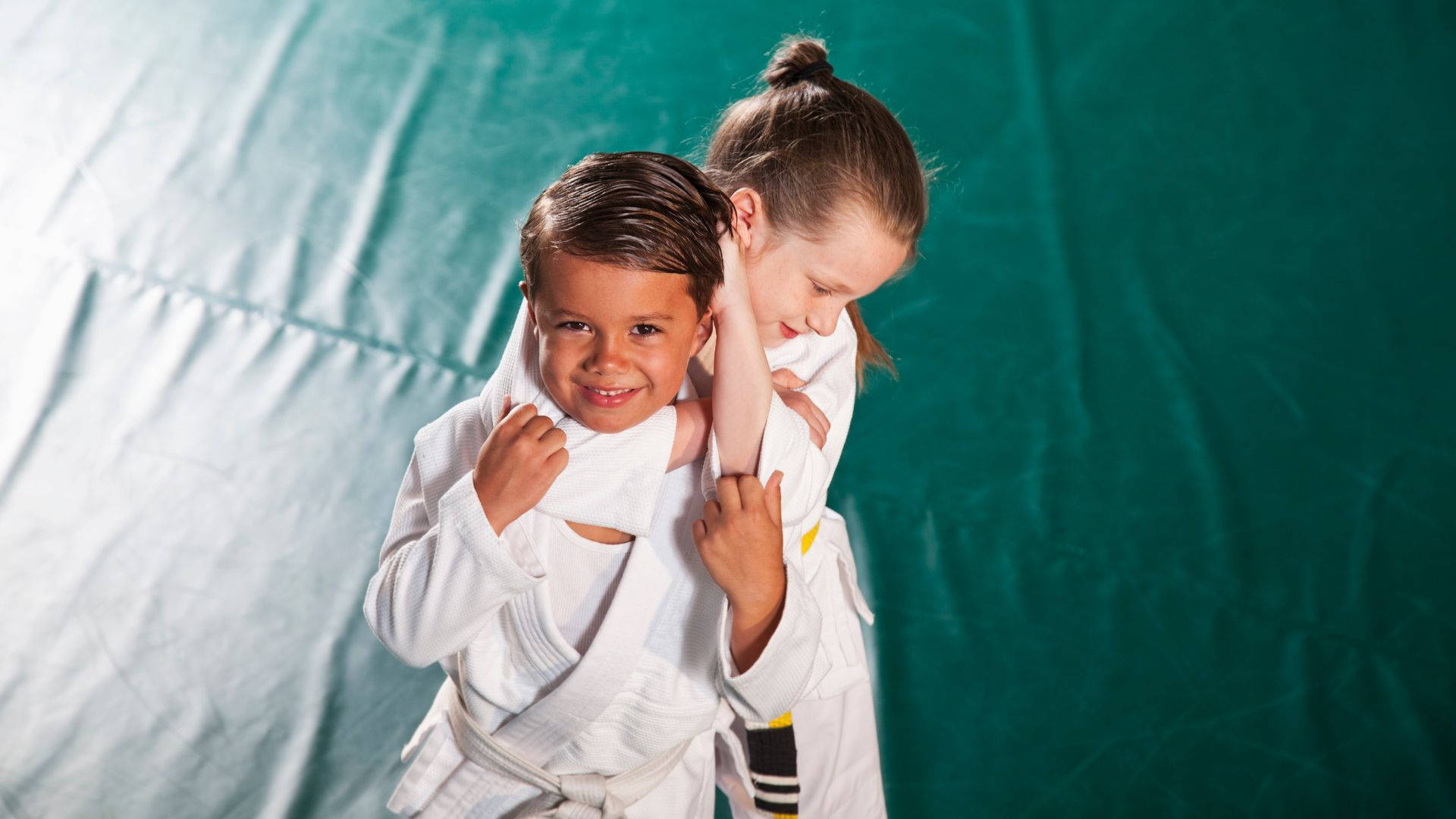martial arts 桌布 壁紙 — 兩個身穿白色柔道服的小孩在青綠色背景前，在明亮的攝影棚燈光下嬉戲地扭打。
