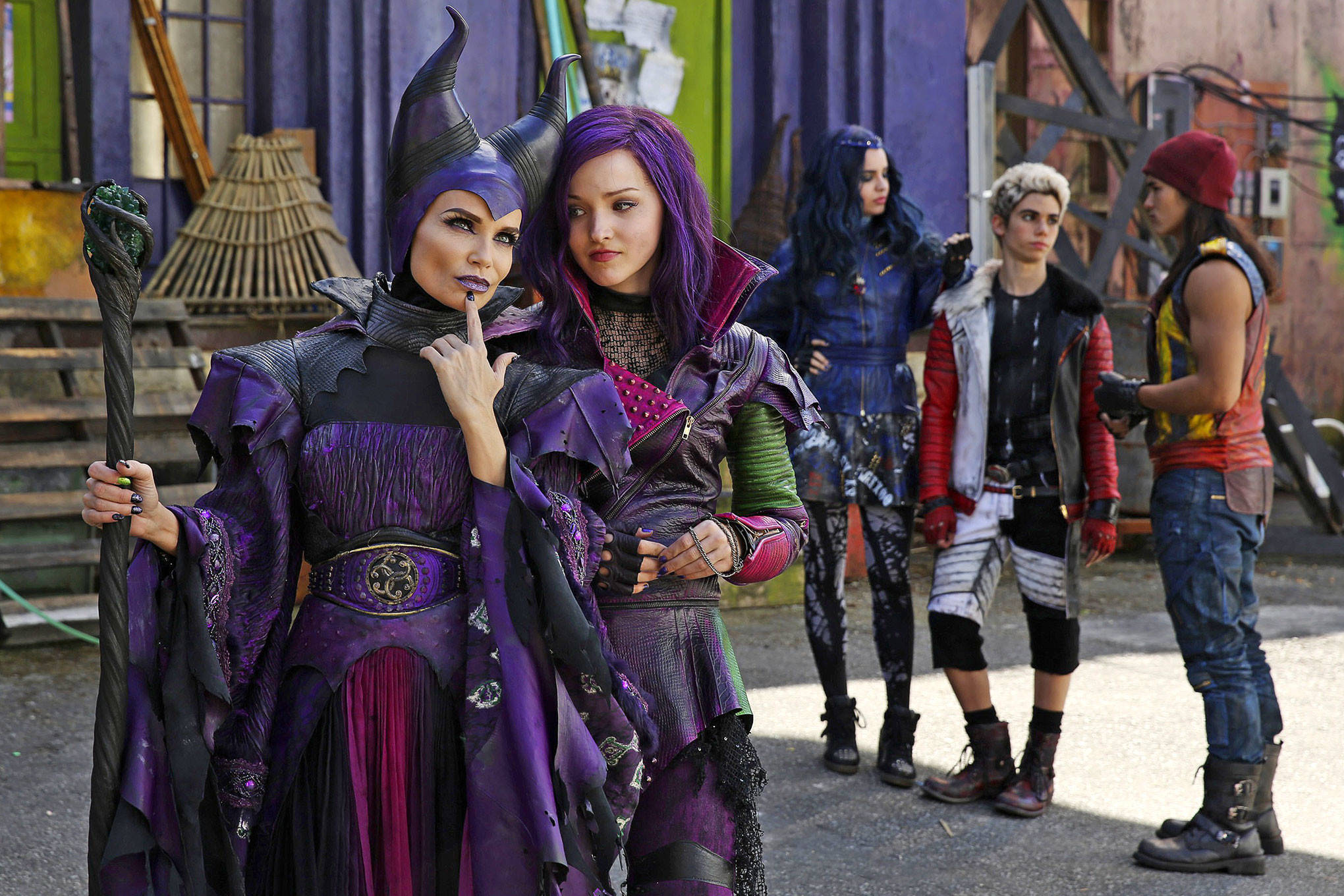 Descendants 桌布 壁紙 — 兩個身穿精緻紫色服裝、頭上有角的巫師站在色彩繽紛的城市背景前，還有另外三個角色。