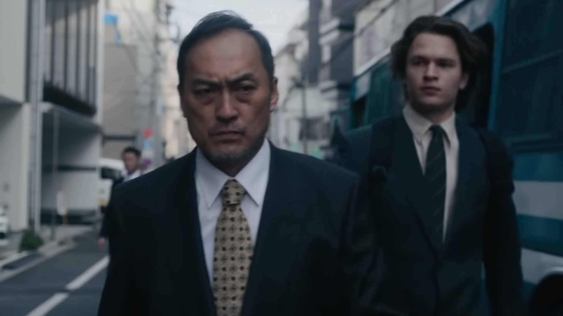 電影動圖 桌布 壁紙 — 兩個身穿西裝的男人在陰天的天空下，帶著嚴肅的表情走過一條狹窄的城市街道。