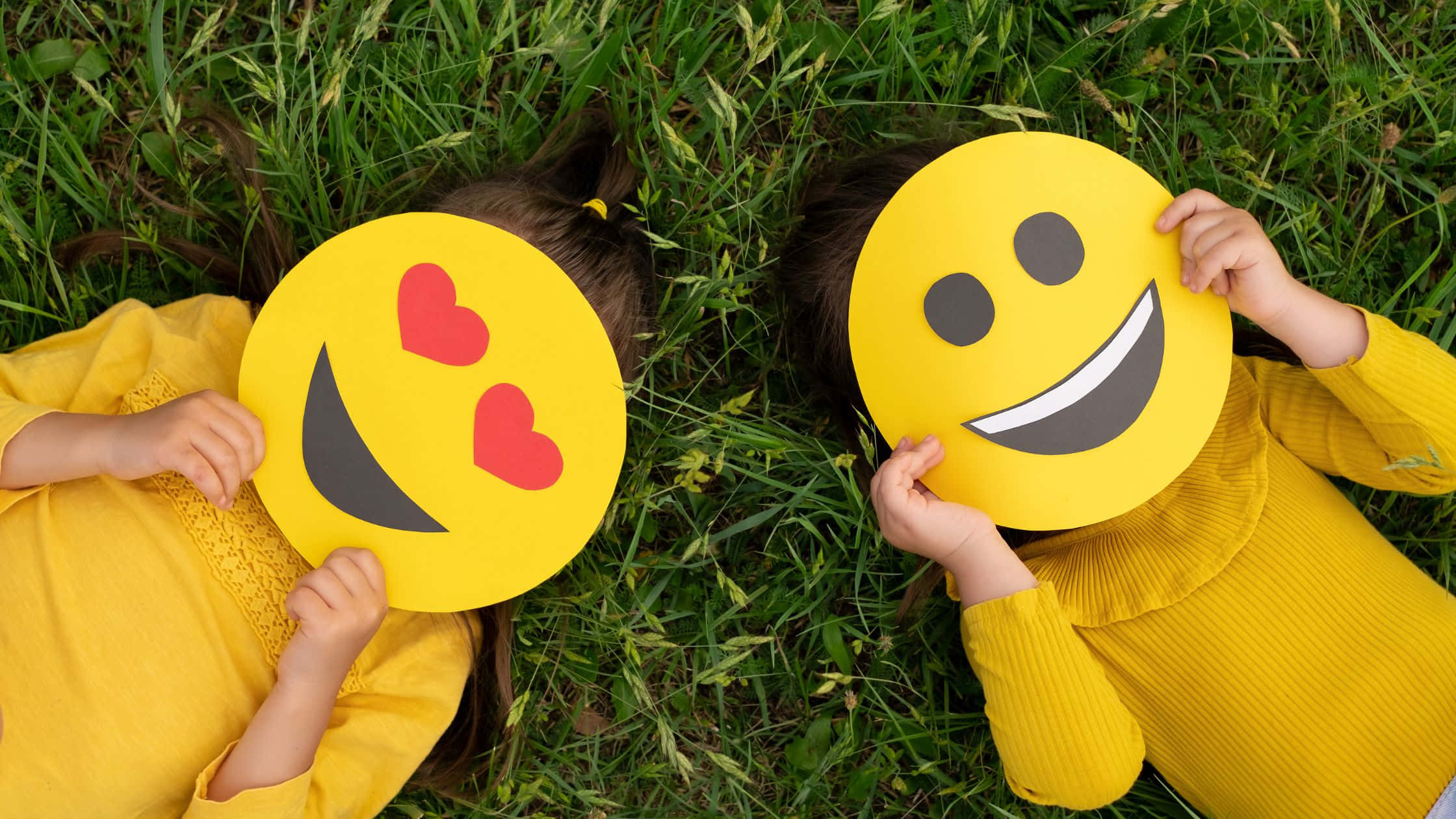 Smile 桌布 壁紙 — 兩個身穿黃色襯衫的孩子躺在草地上，手持超大的表情符號剪紙遮住臉，一個是心形眼睛，另一個是咧嘴笑。