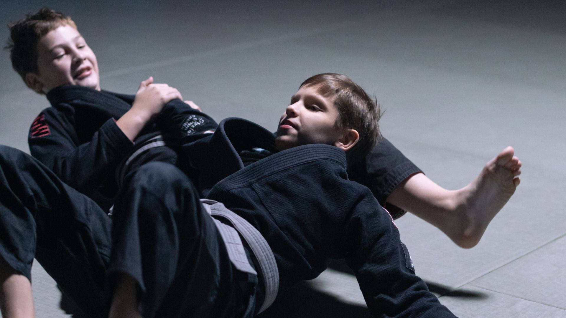 martial arts 桌布 壁紙 — 兩個身穿黑色柔道服的男孩在灰色軟墊上激烈纏鬥，背景是戲劇性的攝影棚燈光。