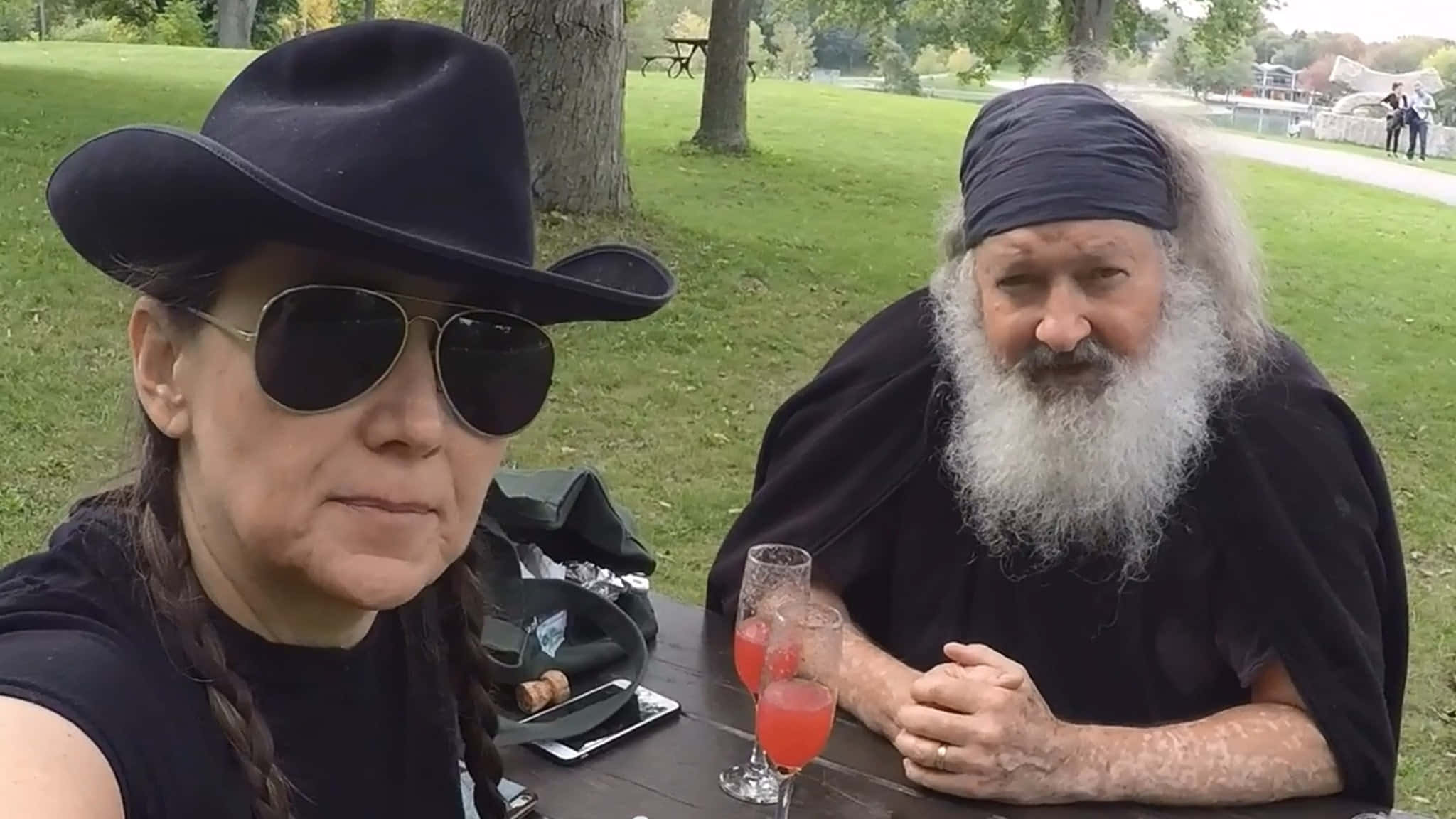 Randy Quaid 桌布 壁紙 — 兩個身穿黑色衣服、戴著帽子的人坐在公園野餐桌旁，在明亮的日光下享用粉紅色飲料。