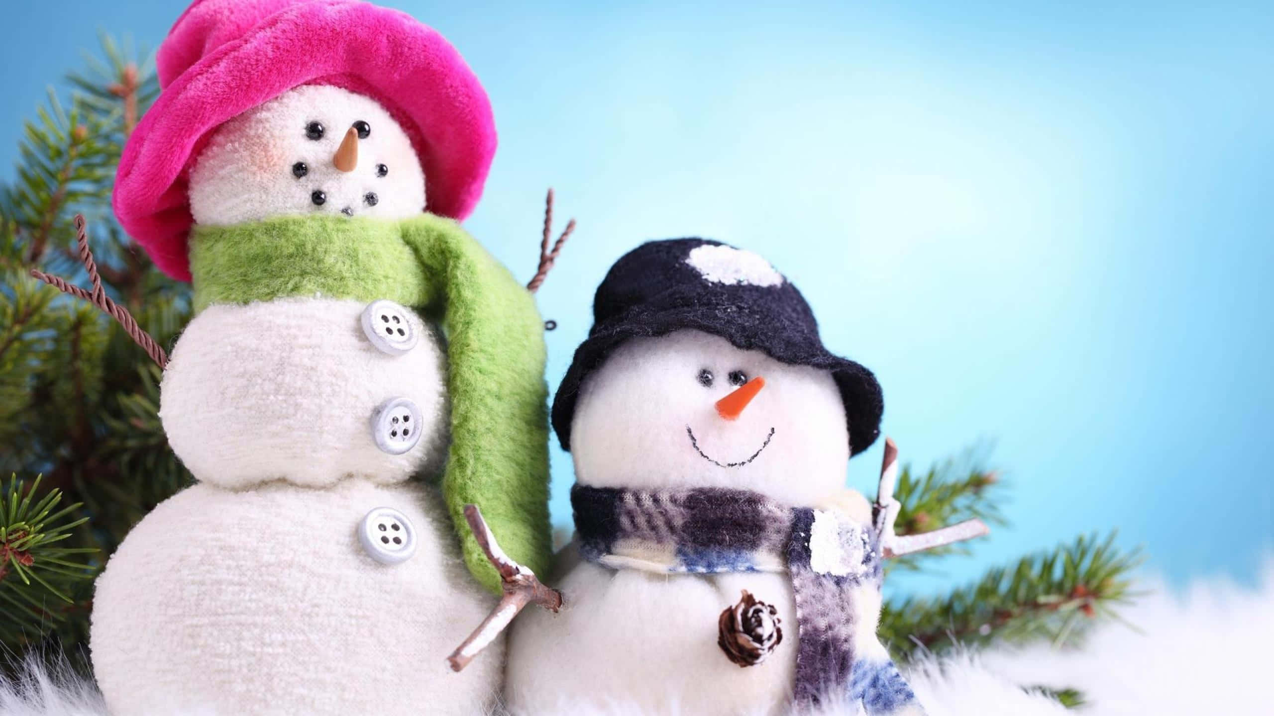 雪 桌布 壁紙 — 兩個迷人的雪人公仔，戴著色彩繽紛的帽子和圍巾，站在松樹枝旁，背景是柔和的藍天。