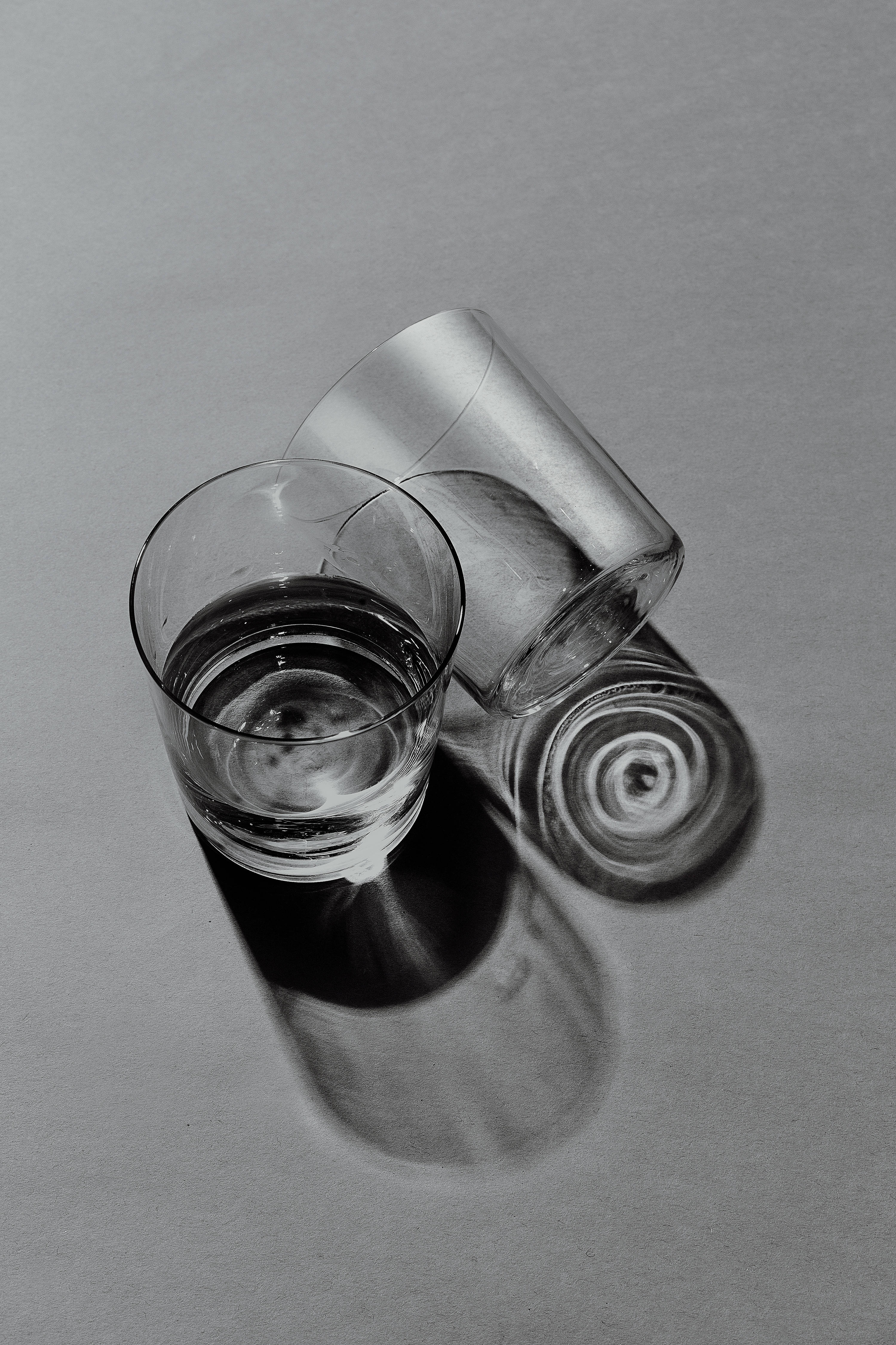 酷 桌布 壁紙 — 兩個透明玻璃杯，一個直立裝水，另一個傾倒，在灰色中性表面上投射出戲劇性的陰影。