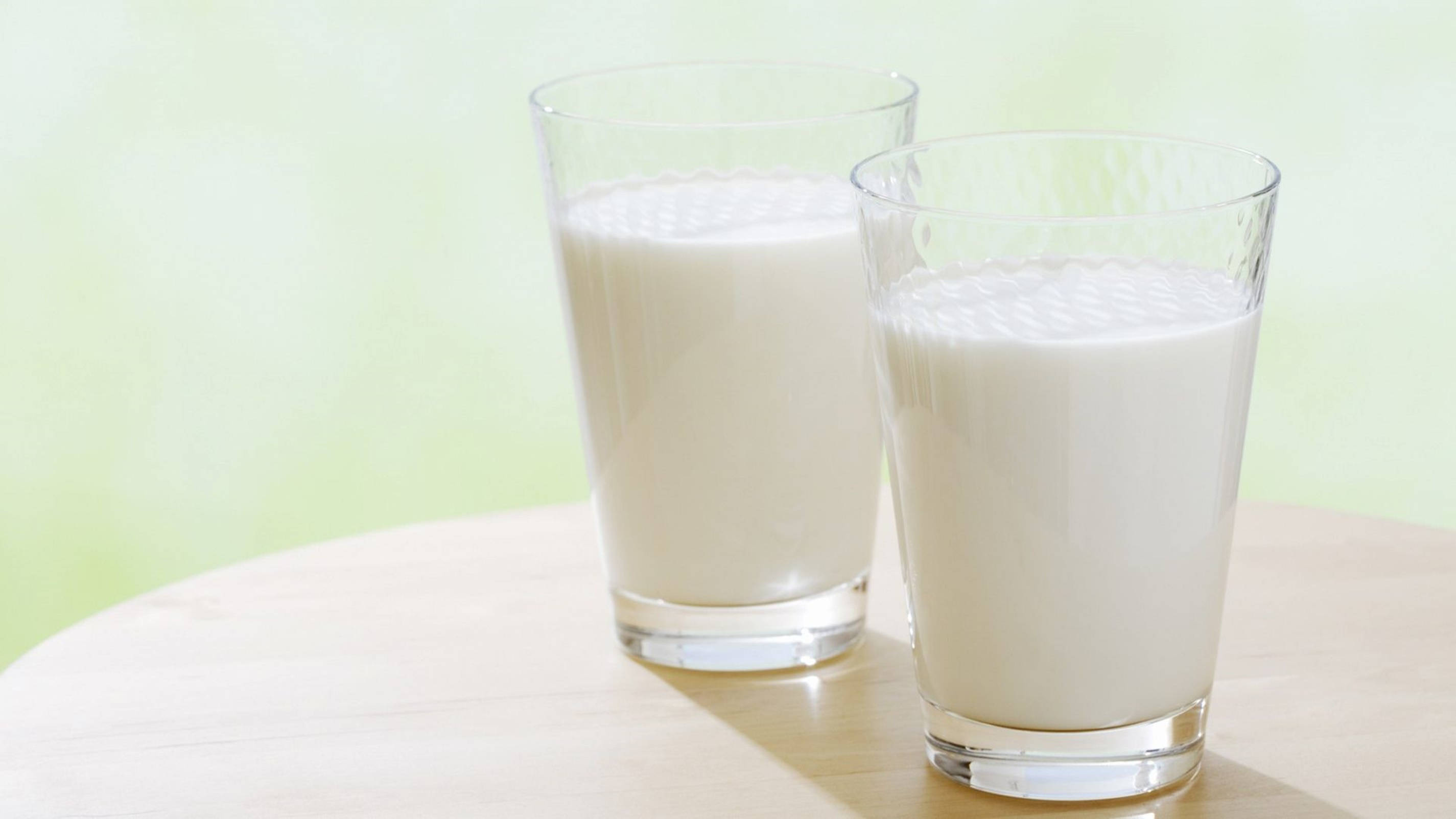 Milk 桌布 壁紙 — 兩個透明玻璃杯裝滿了奶油狀的白色牛奶，放在淺色木桌上，背景是柔和的綠色。