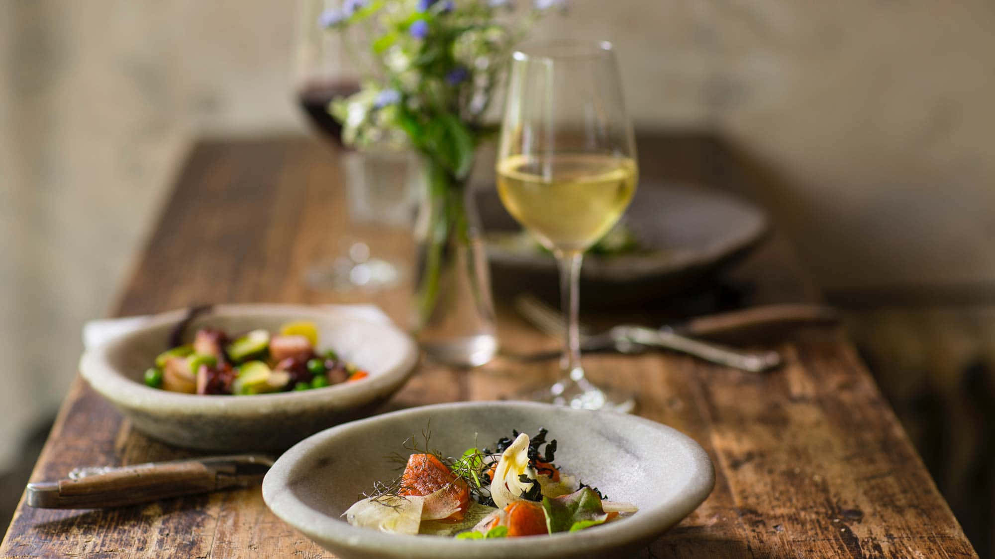 Food And Beverage 桌布 壁紙 — 兩個鄉村風格的碗盛載色彩繽紛的美食沙拉，放在一張經風化的木桌上，旁邊有白酒和紅酒杯，以及一束藍色花朵的花瓶，背景柔和。