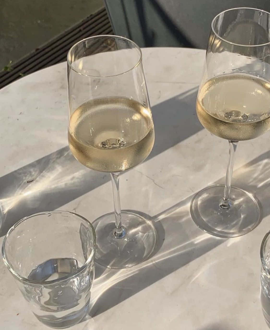 White Wine 桌布 壁紙 — 兩個酒杯和一個水杯放在一張大理石桌上，沐浴在明亮的陽光下，投下清晰的陰影。