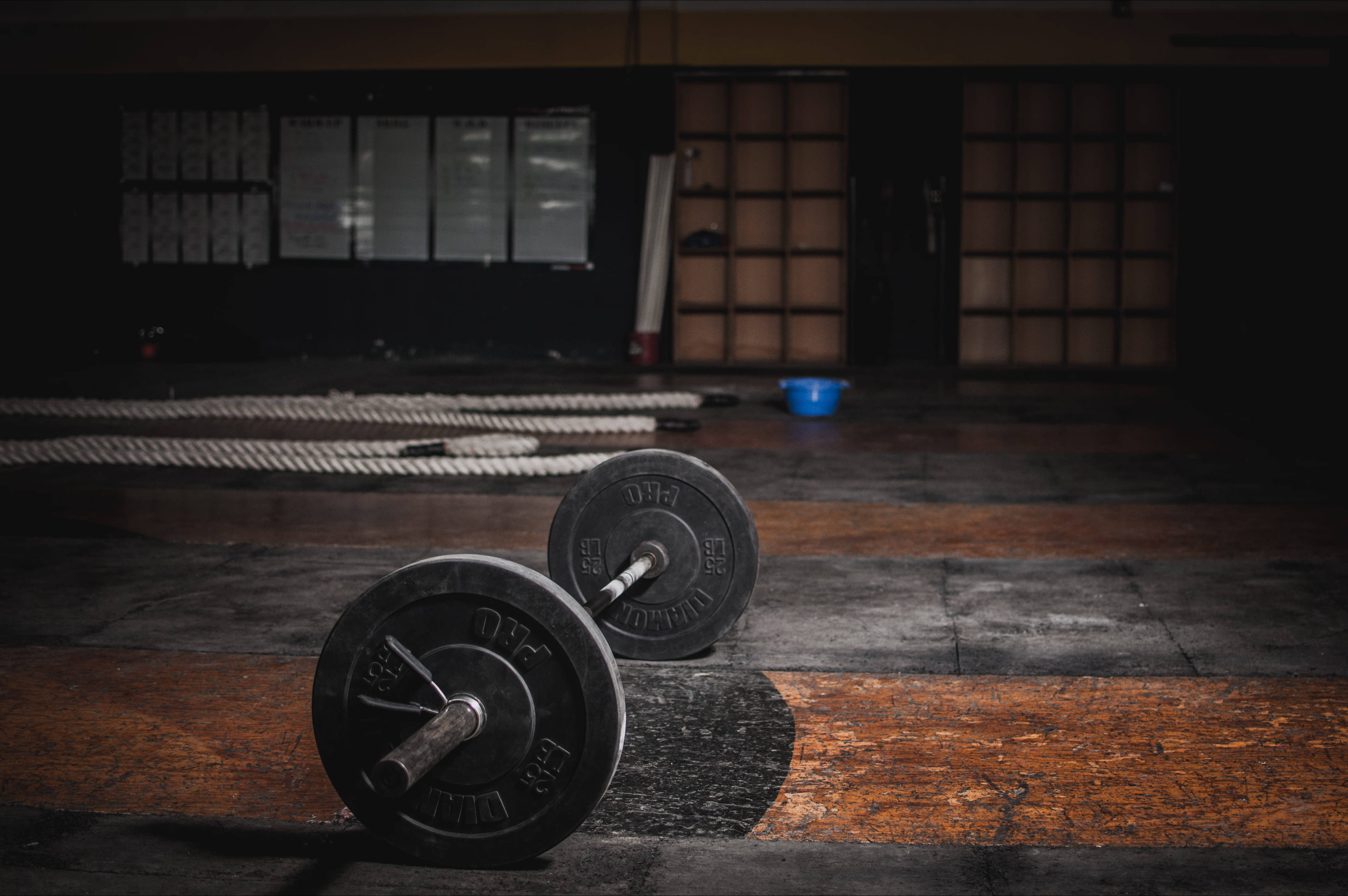 Barbell 桌布 壁紙 — 兩個重型啞鈴放在磨損的木地板上，背景是光線昏暗的健身房，有繩索和障子門。