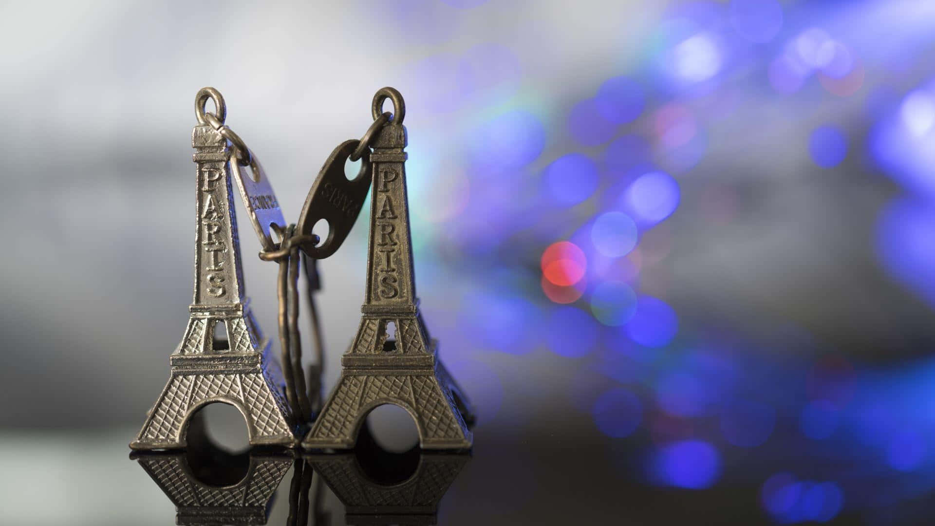 Eiffel Tower 桌布 壁紙 — 兩個金屬製的艾菲爾鐵塔鑰匙圈掛在模糊的藍色散景背景上，帶有柔和的光線反射。