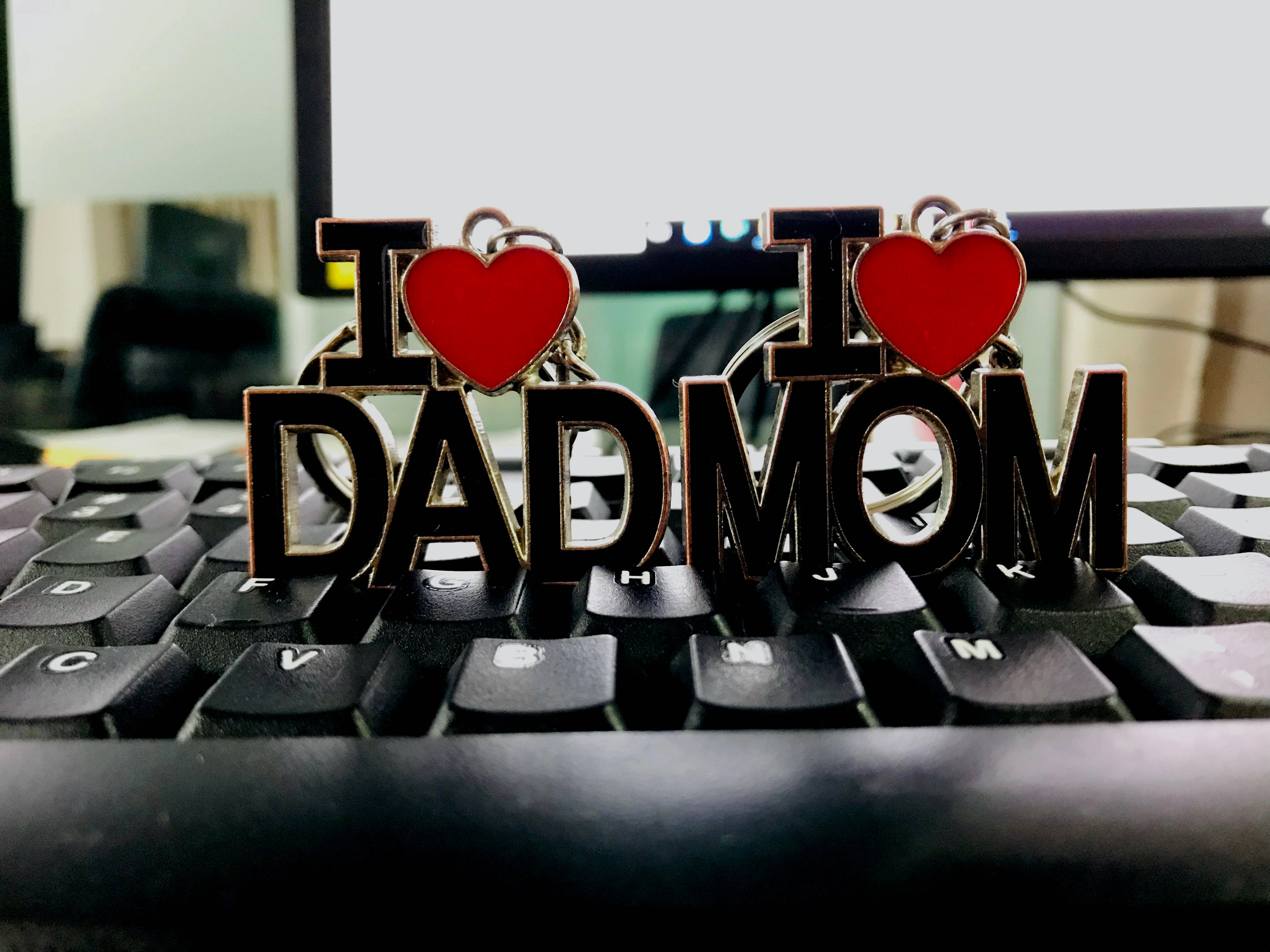Mom 桌布 壁紙 — 兩個金屬鑰匙圈，分別拼寫 I heart DAD 和 I heart MOM，放在一個黑色電腦鍵盤上，背景是模糊的辦公室。