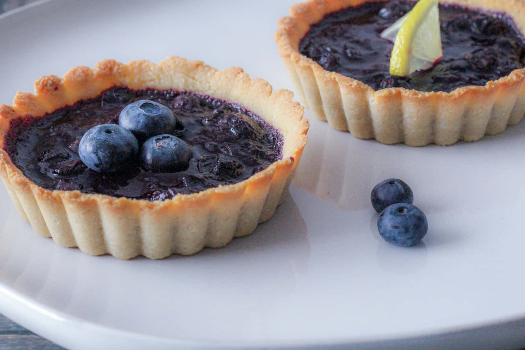 Blueberries Tart 桌布 壁紙 — 兩個金色藍莓塔，帶有光澤的餡料，上面放上新鮮莓果和檸檬角，放在白色盤子上。