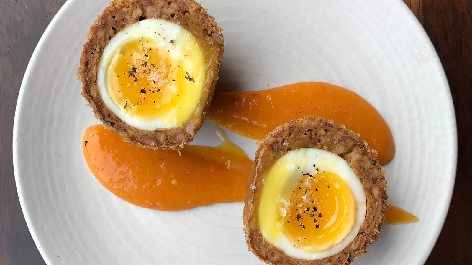 Egg 桌布 壁紙 — 兩個金黃色的蘇格蘭蛋，流心蛋黃配酥脆外皮，放在白色盤子上，旁邊是鮮豔的橙色醬汁。