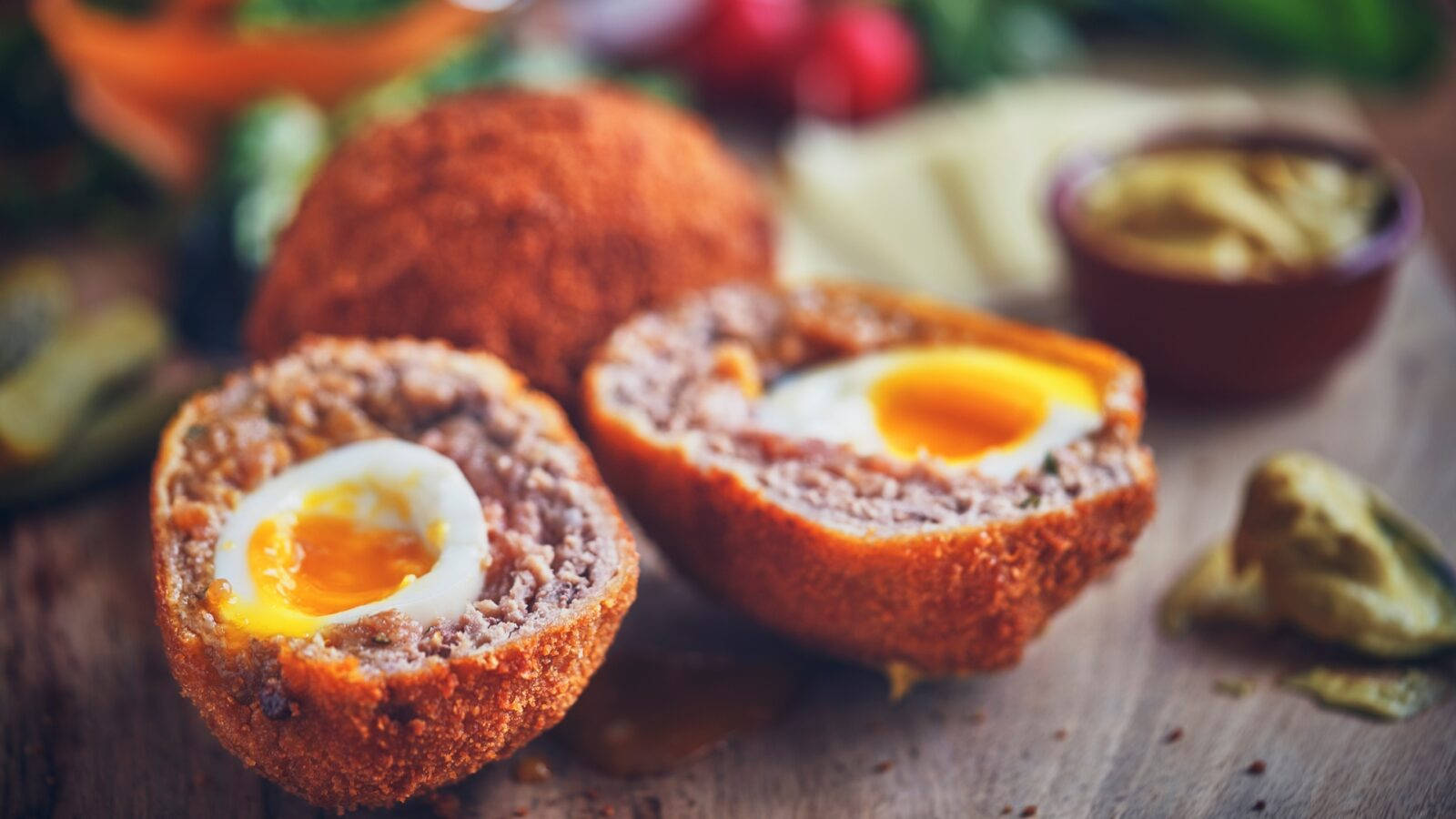 Egg 桌布 壁紙 — 兩個金黃色的蘇格蘭蛋切開，露出流心蛋黃和美味的肉餡，放在質樸的木板上。