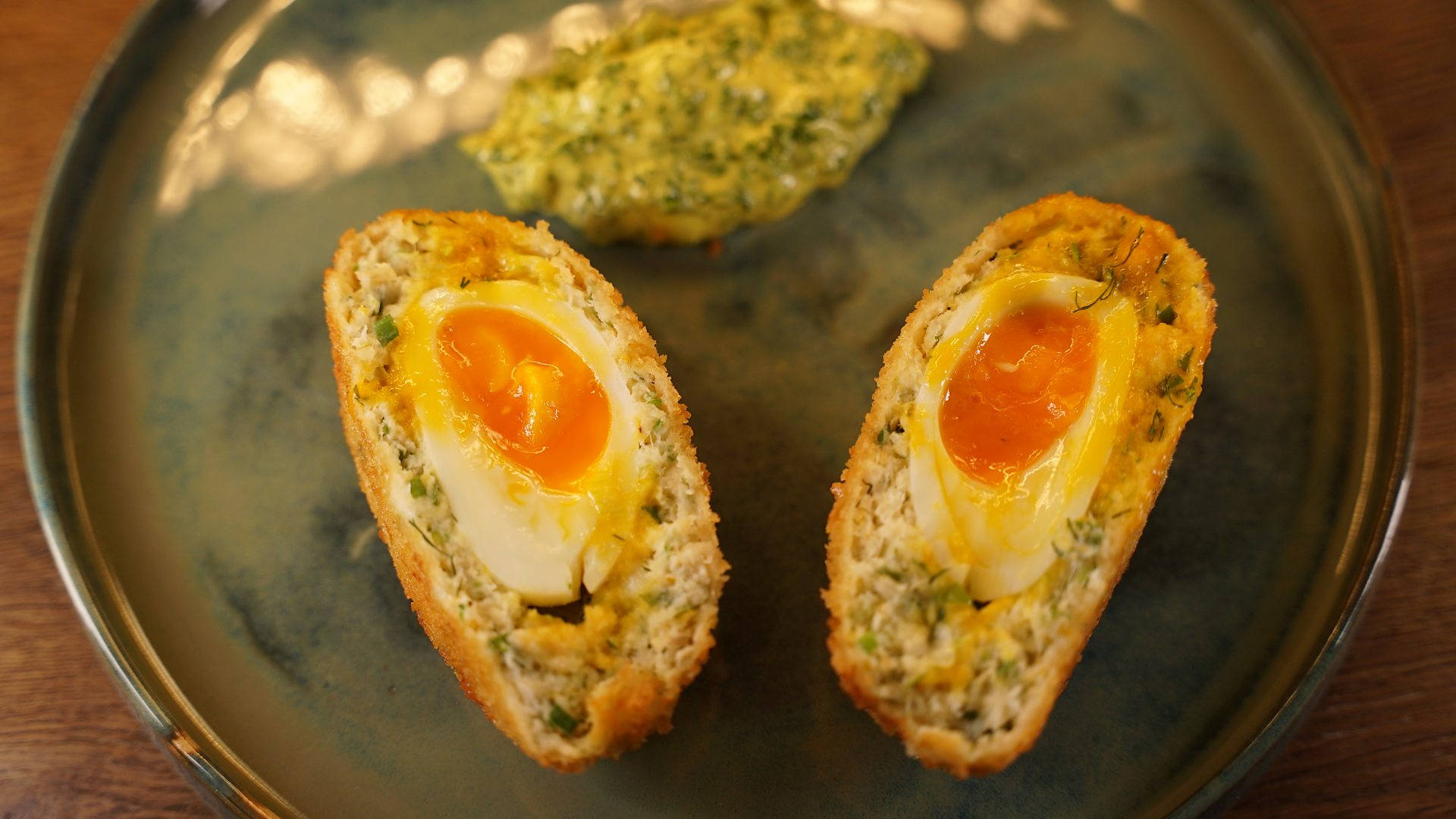 Egg 桌布 壁紙 — 兩個金黃色的蘇格蘭蛋切開，露出流心蛋黃和香草點綴的外皮，放在深色陶瓷盤上。