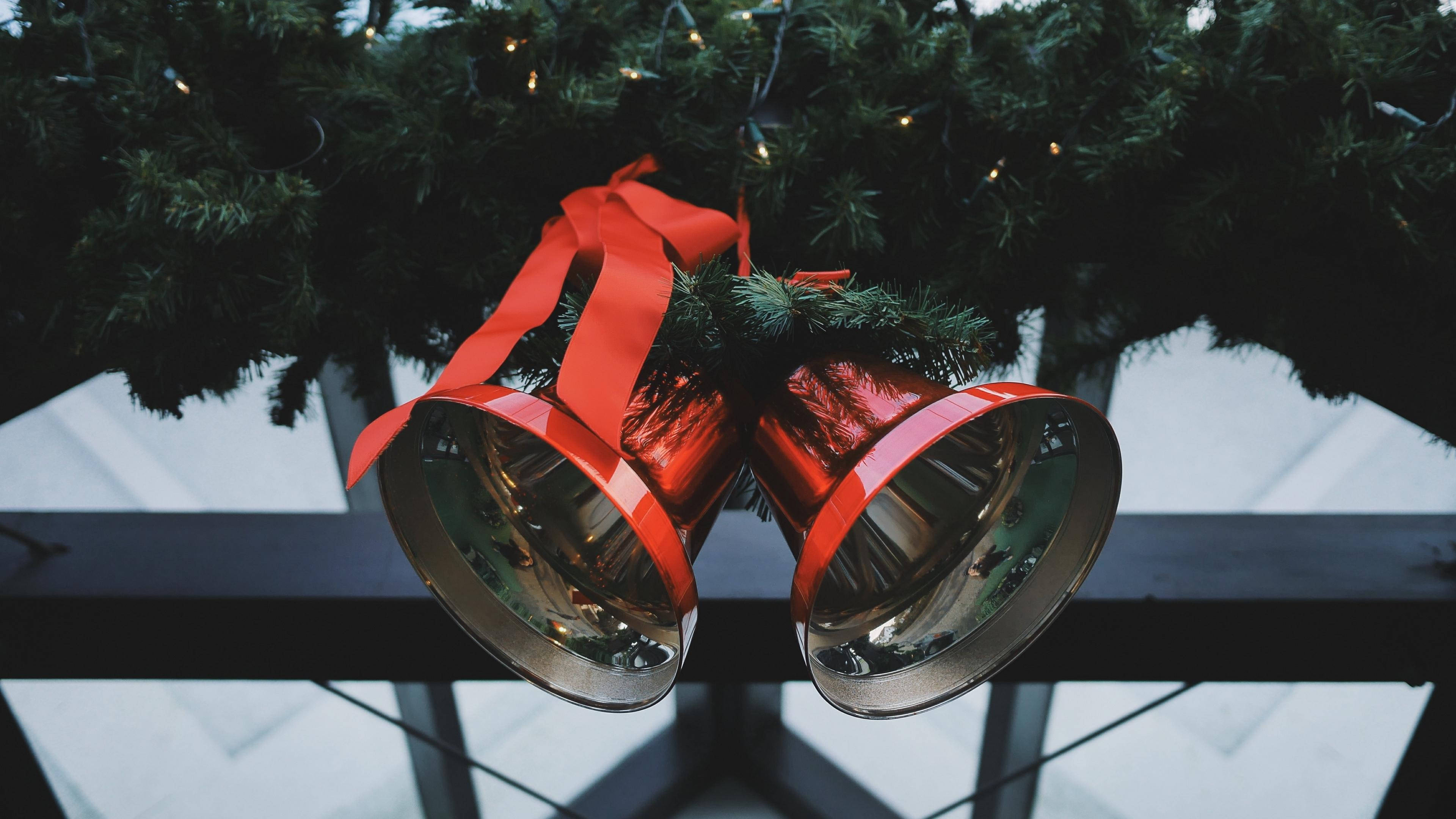 Christmas Bell 桌布 壁紙 — 兩個閃亮的銀色鈴鐺用紅色絲帶綁著，掛在一條深綠色松樹枝上，枝上還有閃爍的仙女燈。