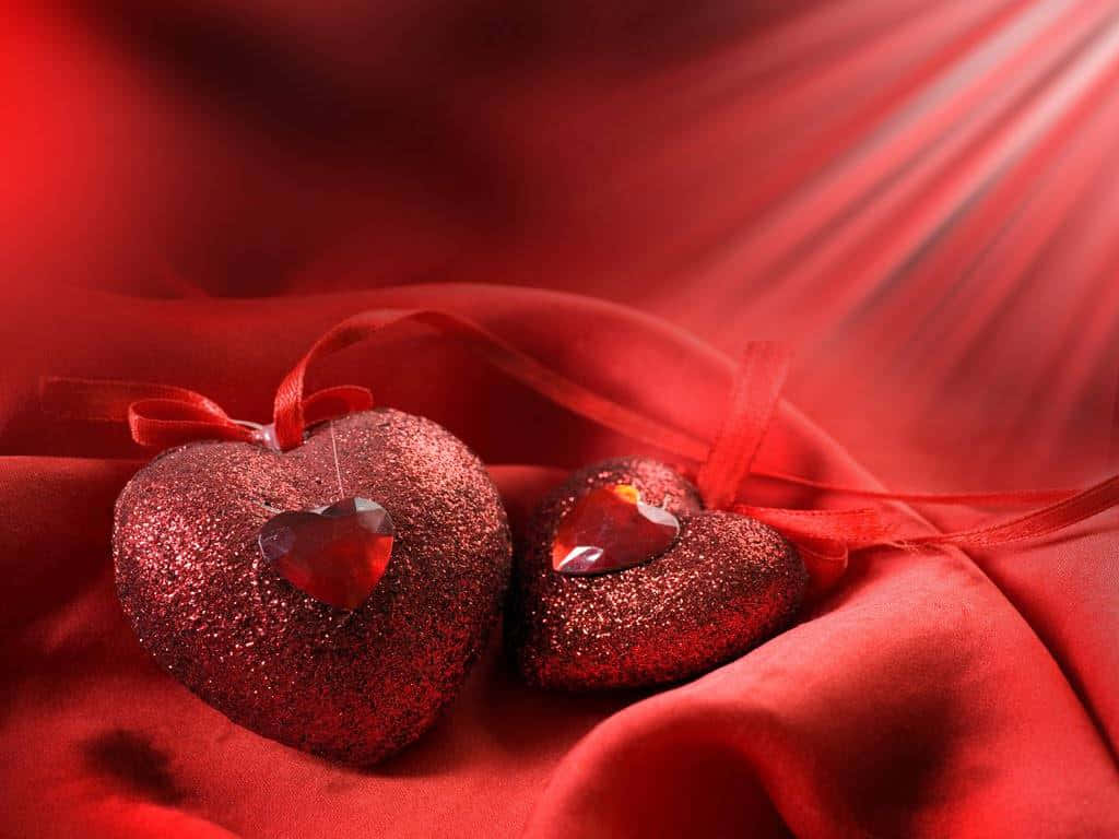 Sweet Love 桌布 壁紙 — 兩個閃閃發光的紅色心形裝飾品，帶有水晶中心，放置在溫暖光線沐浴下的流動紅色布料上。