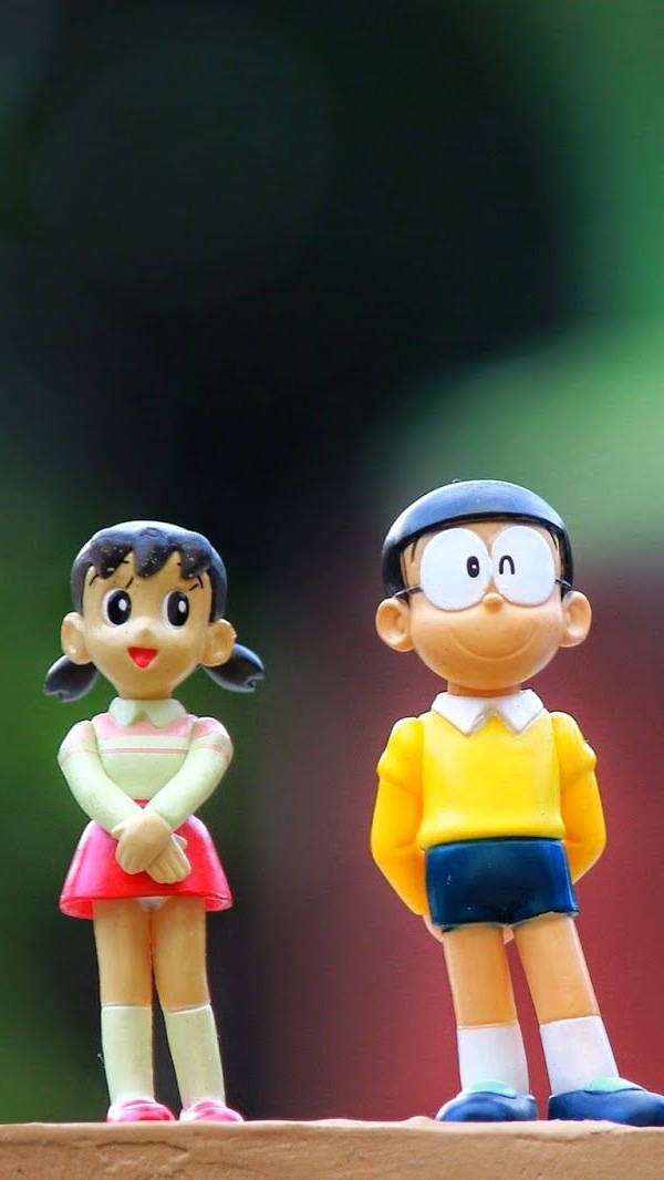 Doraemon 桌布 壁紙 — 兩個開心的動漫公仔哆啦A夢和靜香並排站在木質表面上，背景是模糊的綠色和紅色。
