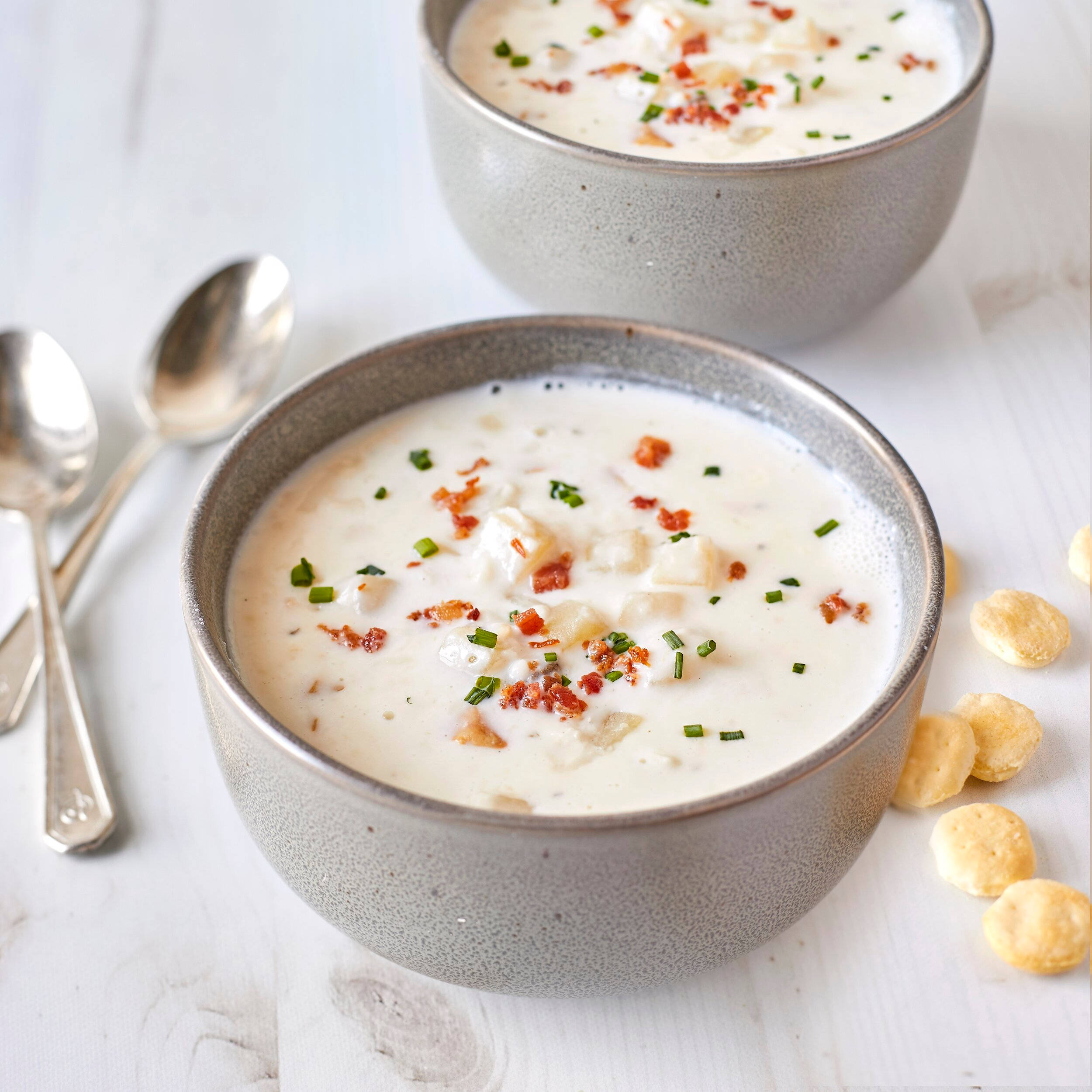 Soup 桌布 壁紙 — 兩個陶瓷碗裝著奶油蛤蜊湯，上面放著脆培根和切碎的細香蔥，旁邊有蘇打餅乾和湯匙，放在白色木桌上。